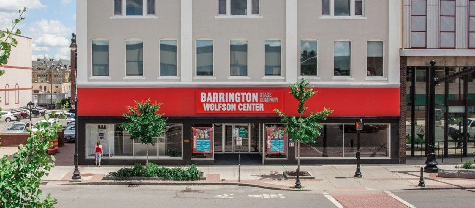 Street view of a building with a red sign that reads 'Barrington Wolfson Center Stage Company' above the entrance. There are two small trees, street lamps, and parked cars visible.