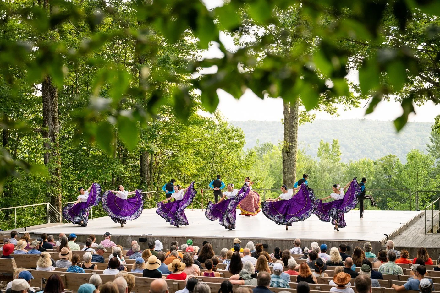 A group of dancers in traditional Mexican dresses performing on an outdoor stage with a forest and mountains in the background, while an audience watches.