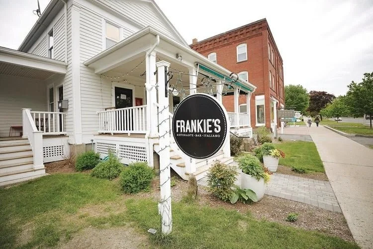 White building with outdoor patio, sign for Frankie's Italian restaurant, sidewalk, and adjacent red brick building