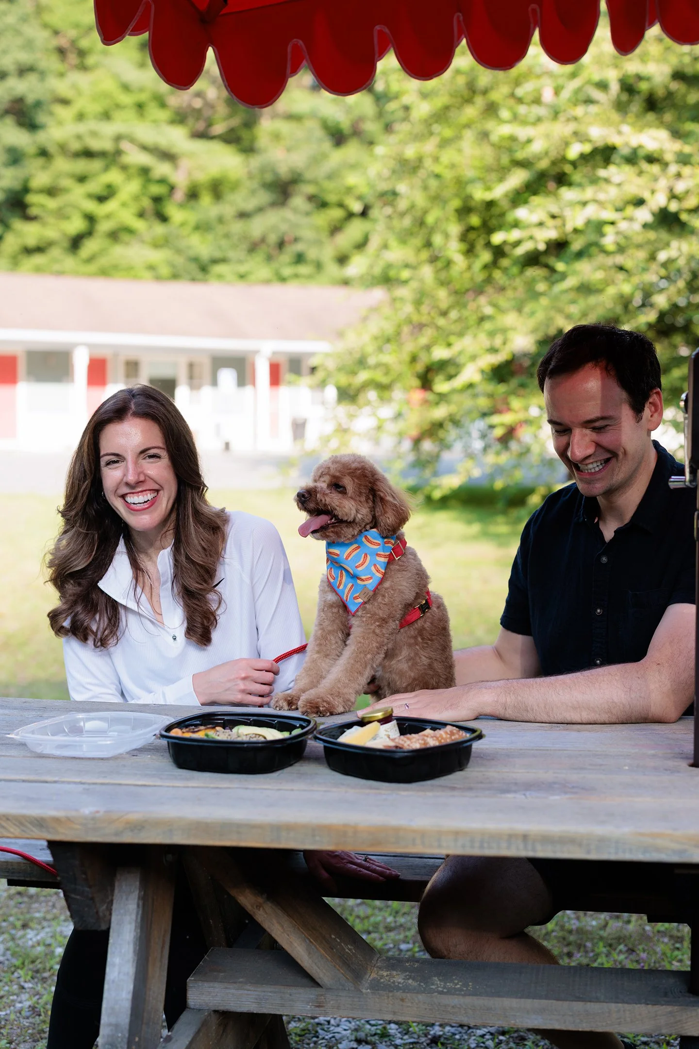 A man and a woman sitting at an outdoor picnic table with food containers and a small dog between them. The dog is wearing a blue bandana with fish pattern. The woman has long brown hair and is laughing, while the man with short dark hair is smiling. There is greenery and a house in the background.