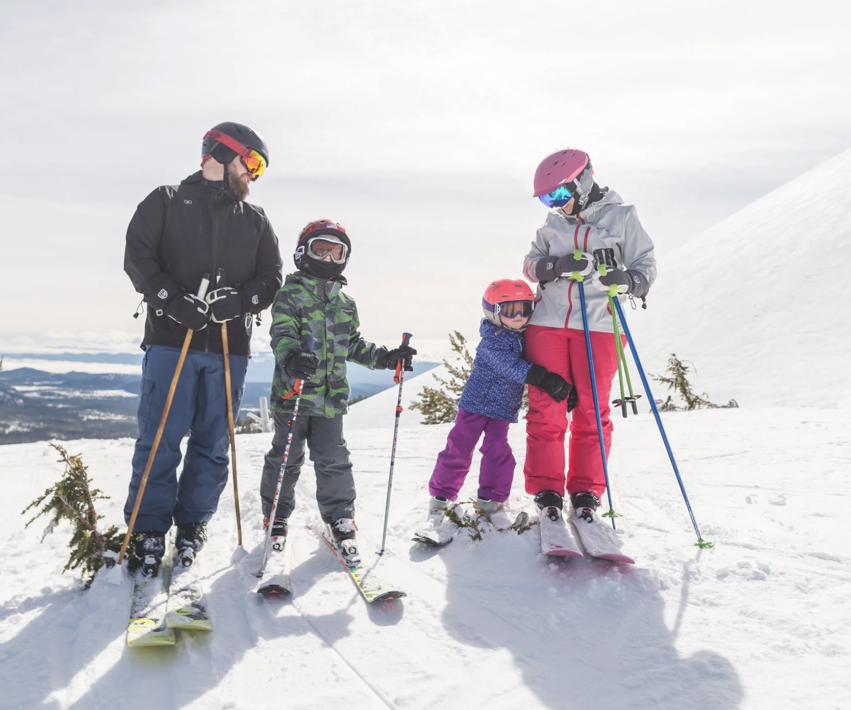 A family of five standing on snow with skis and poles, dressed in winter gear including helmets and goggles, on a snowy mountain with a cloudy sky.