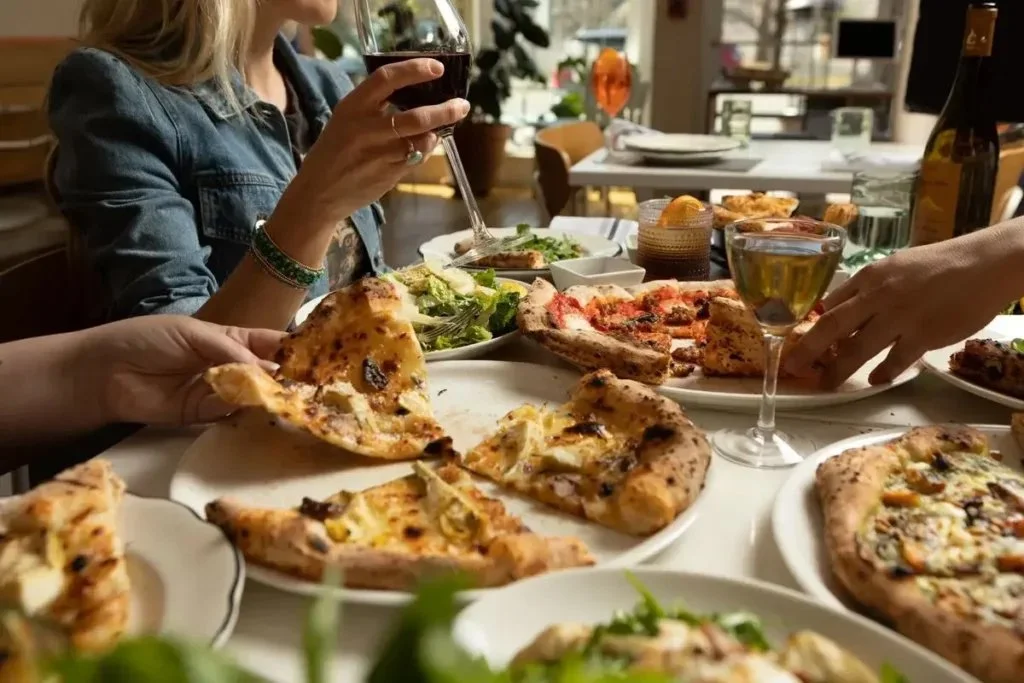 People enjoying a meal with multiple plates of pizza, salad, and drinks on a table at a restaurant.