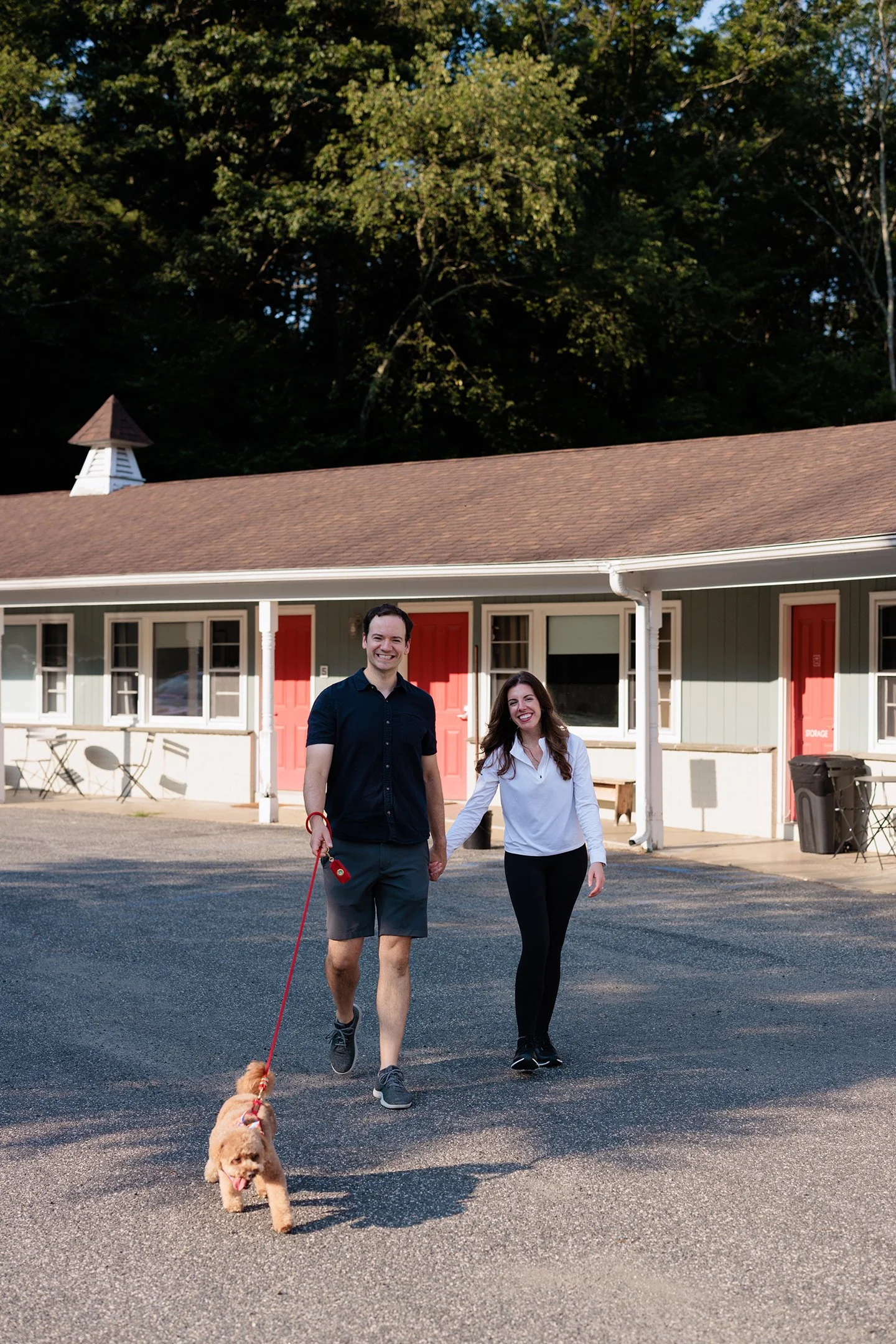 A young man and woman walk hand in hand on a paved street, smiling, with the man holding a small dog on a leash. They are in front of a row of houses with red doors and a white and green exterior, with a large, leafy tree in the background.