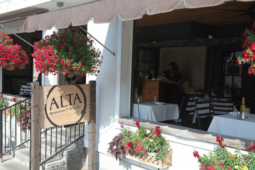 Exterior view of ALTA restaurant and wine bar with hanging flower baskets and a flower box on the windowsill, showing an open window with tables inside and staff preparing inside.