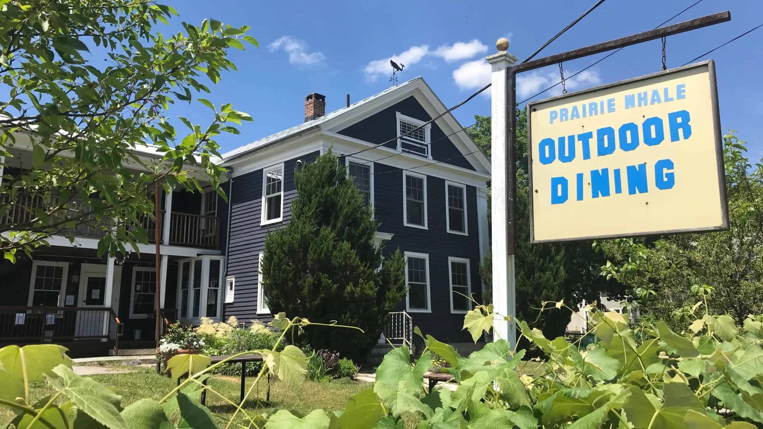 A large dark blue house with white trim, multiple windows, and a porch, surrounded by green trees and plants under a blue sky. A sign reads 'Prairie Whale Outdoor Dining'.