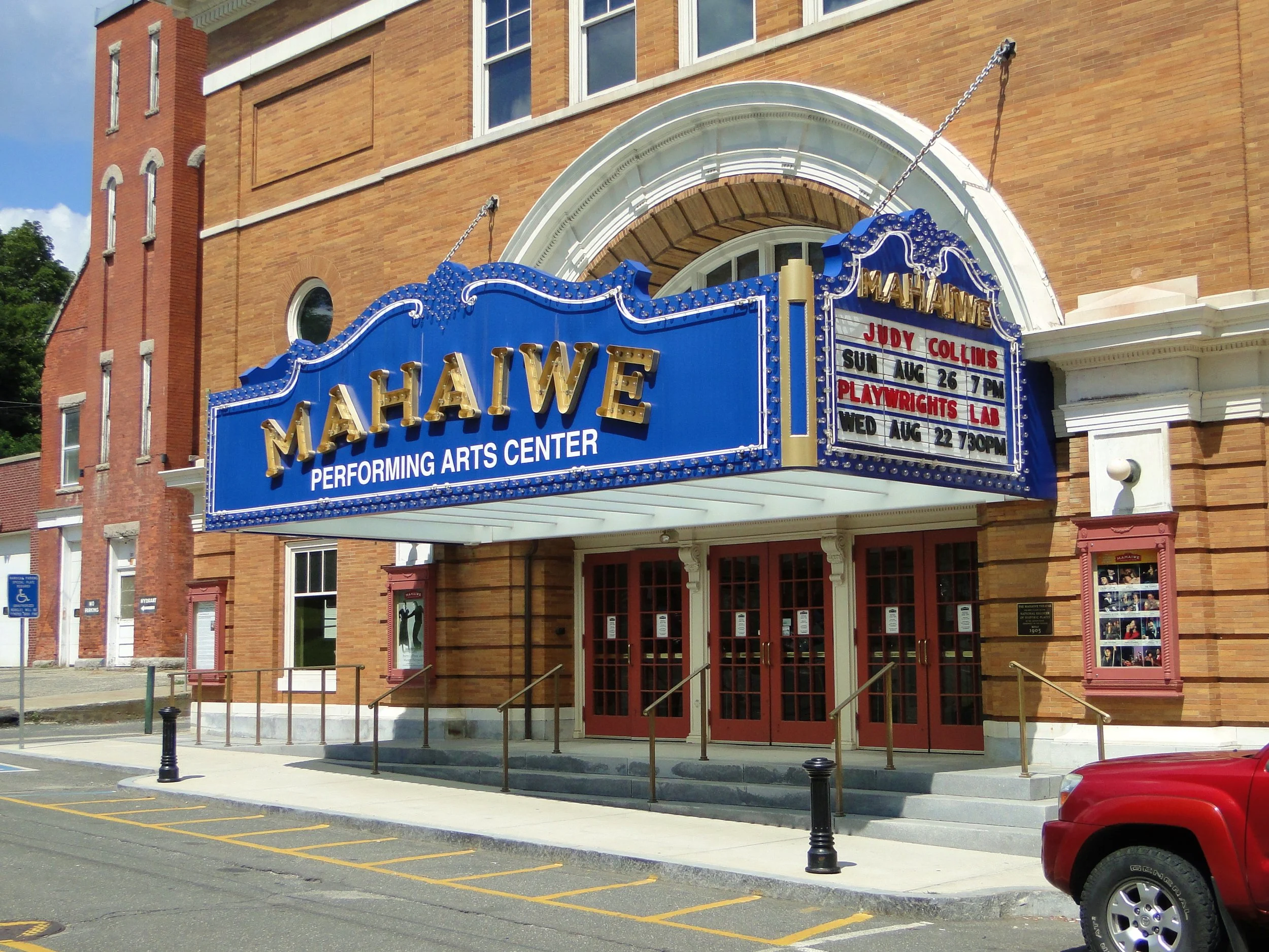 The Maramek Performing Arts Center sign out front of a brick building with red doors, display cases, and large windows. The marquee displays upcoming event information.