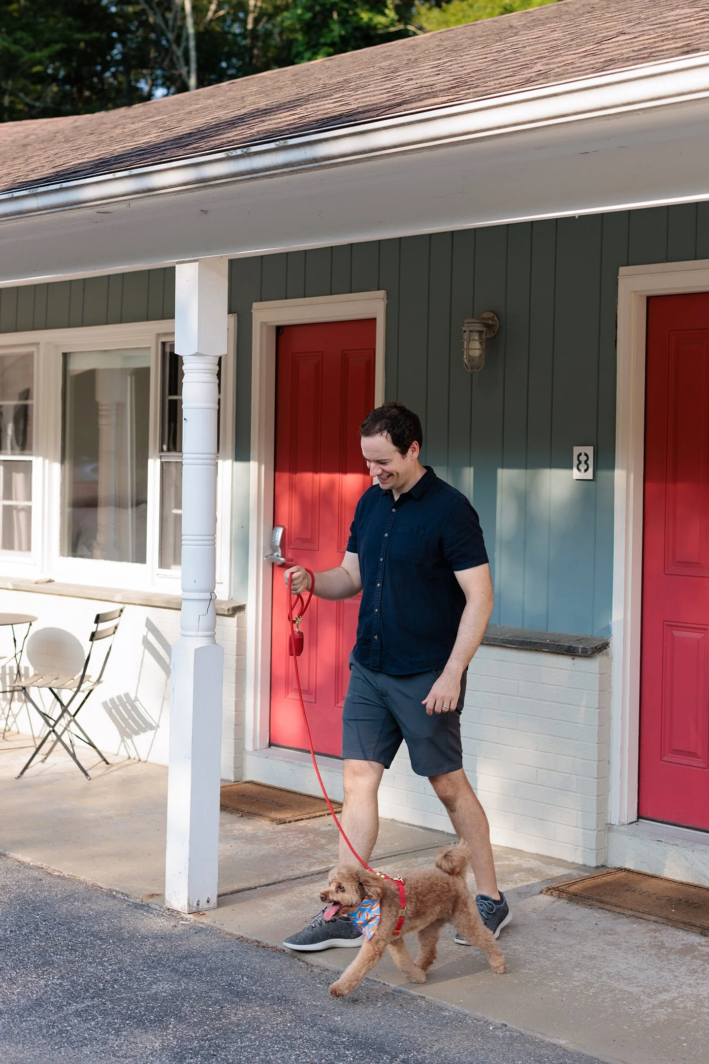 A man walking a small dog on a leash outside a house with red doors and green siding. The man is smiling and wearing a navy shirt and shorts, and the dog has a colorful bandana.