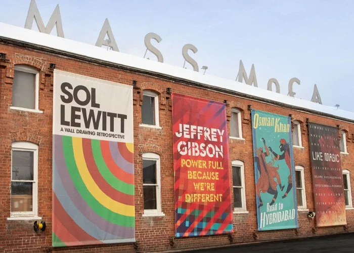 Four colorful banners hung on a brick building with the partially visible word 'MASS MoCA' on the rooftop.