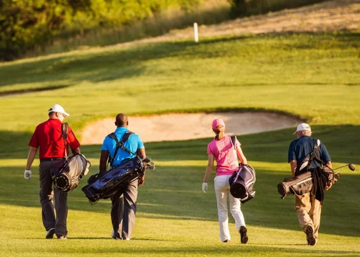 Four golfers walking on the golf course with golf bags, approaching a green and sand trap, during a sunny day.