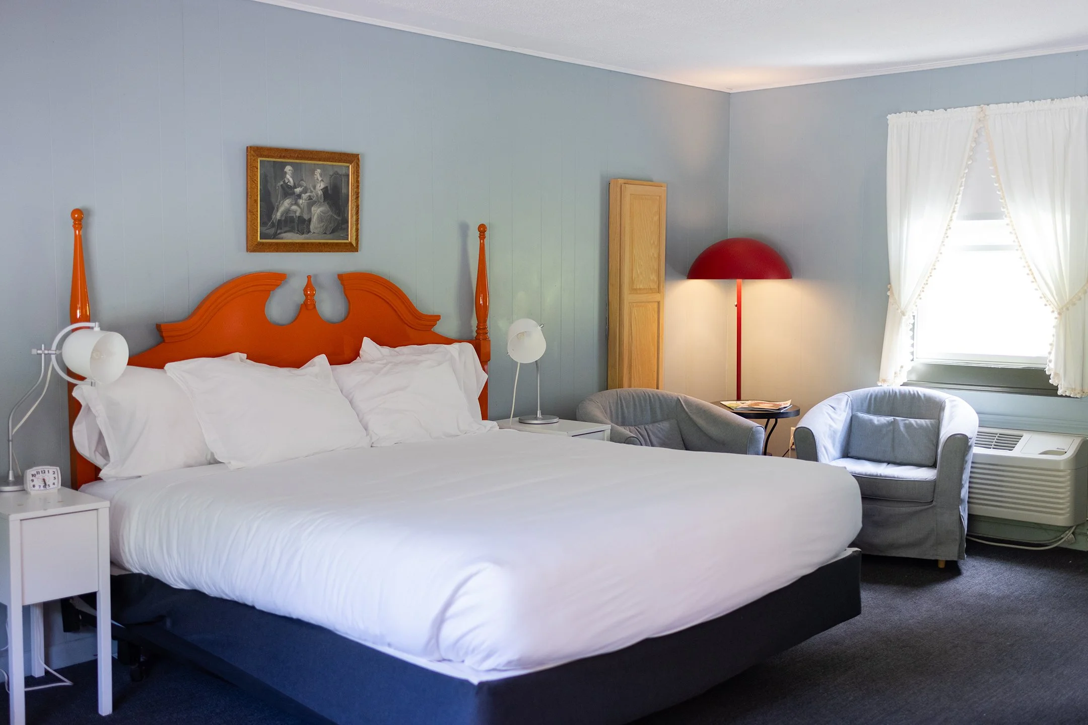 A bedroom with a bed featuring an orange headboard, white bedding, and pillows. There are two bedside tables, one with a small clock. A framed picture hangs above the bed. To the right, there is a standing red lamp, a small closet, and a window with white curtains. Two gray armchairs and a small table with magazines are near the window, with an air conditioning unit below.