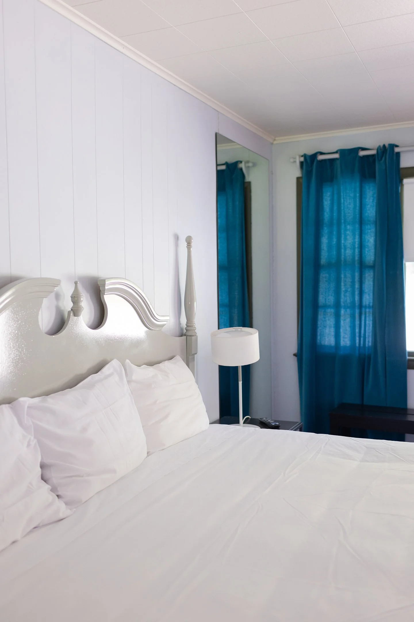 A bedroom with a bed covered in white bedding and pillows, a white nightstand with a lamp, a mirror on the wall, and blue curtains over a window.