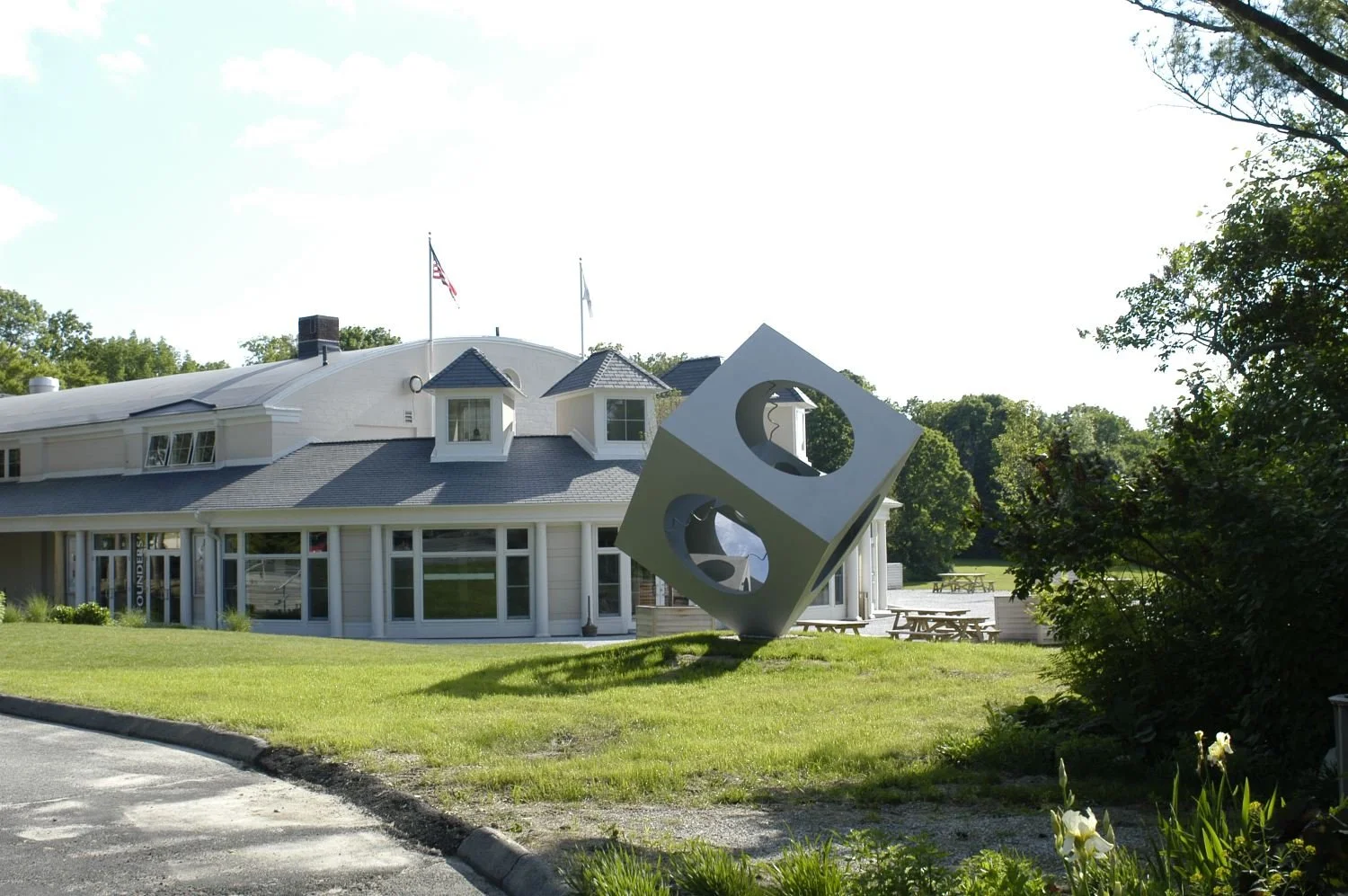 A modern house with a curved roof and multiple windows, with a large white geometric sculpture with circular cutouts in the yard, surrounded by green grass and trees.