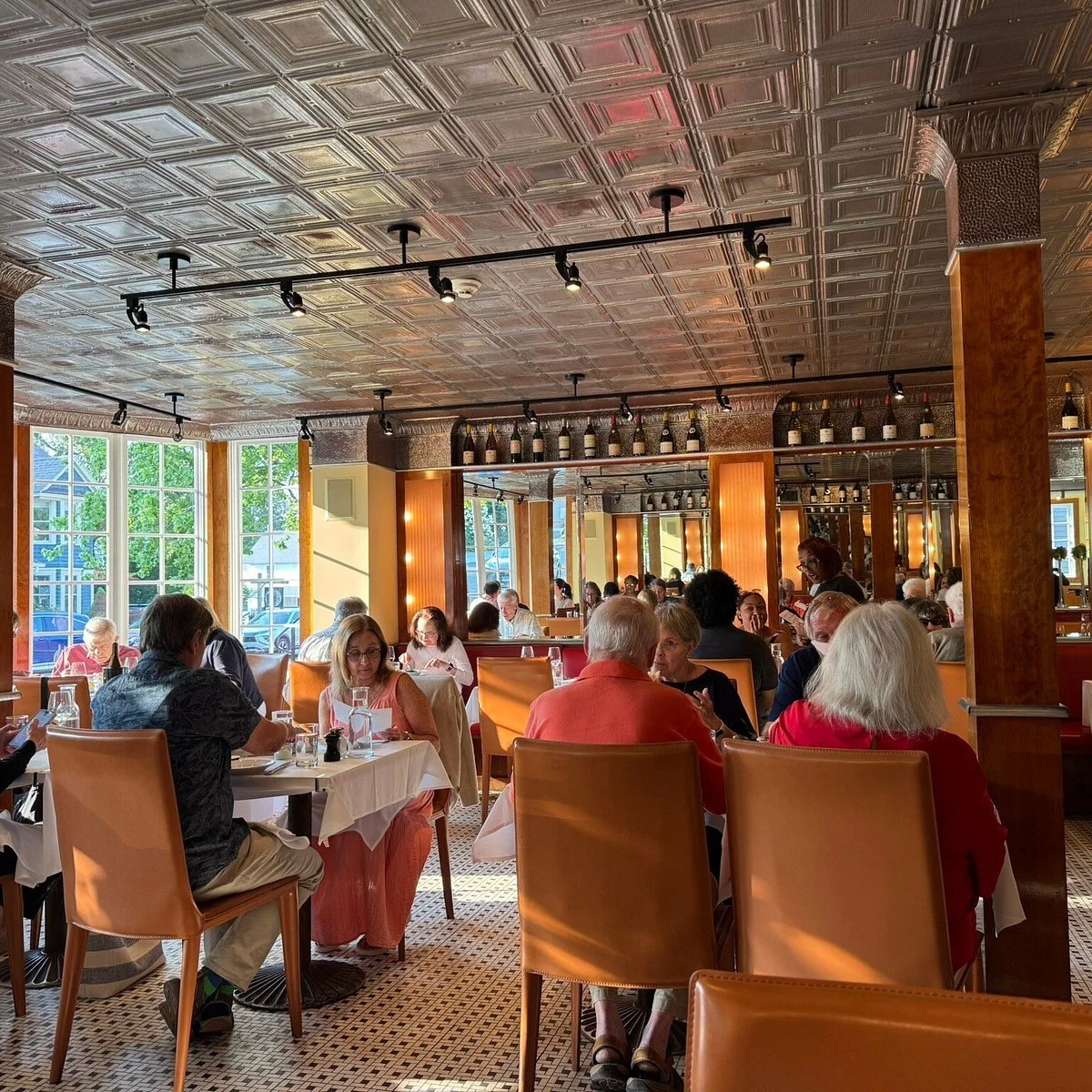 Interior of a restaurant filled with people dining at tables with white tablecloths, large windows letting in natural light, decorative ceiling panels, and bottles of wine on shelves along the wall.