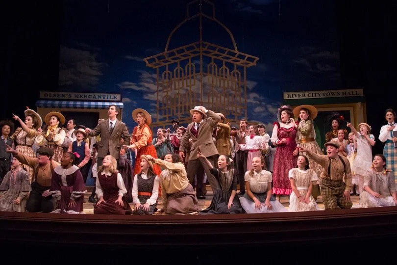 The cast of a theatrical performance, dressed in period costumes, performing on stage with a cityscape backdrop, including signs for Olsens' Mercantile and River City Town Hall.