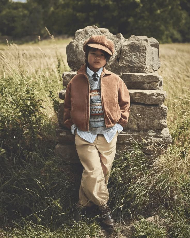 A young boy standing outdoors in front of a stone wall, wearing vintage-style clothing including a brown cap, brown jacket, sweater, shirt, beige pants, and boots, with grassy field and trees in the background.