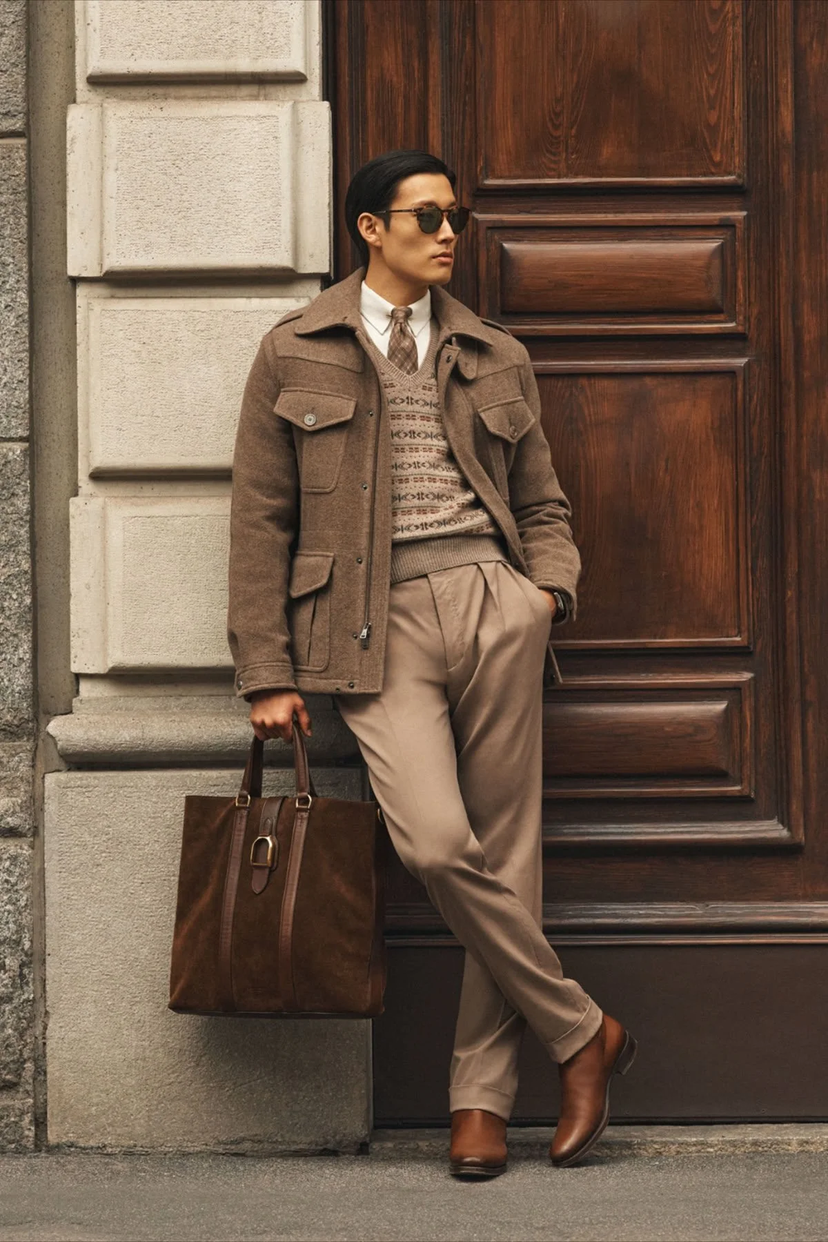 A stylish man with dark hair and sunglasses wearing a brown jacket, patterned sweater, beige trousers, and brown boots, leaning against a wooden door while holding a brown leather bag.