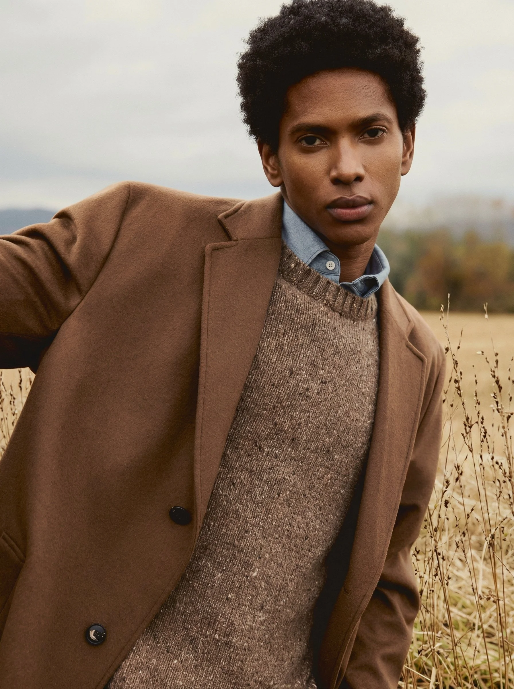 A young man with dark curly hair standing in a field of tall, dry grass during daytime with a cloudy sky.
