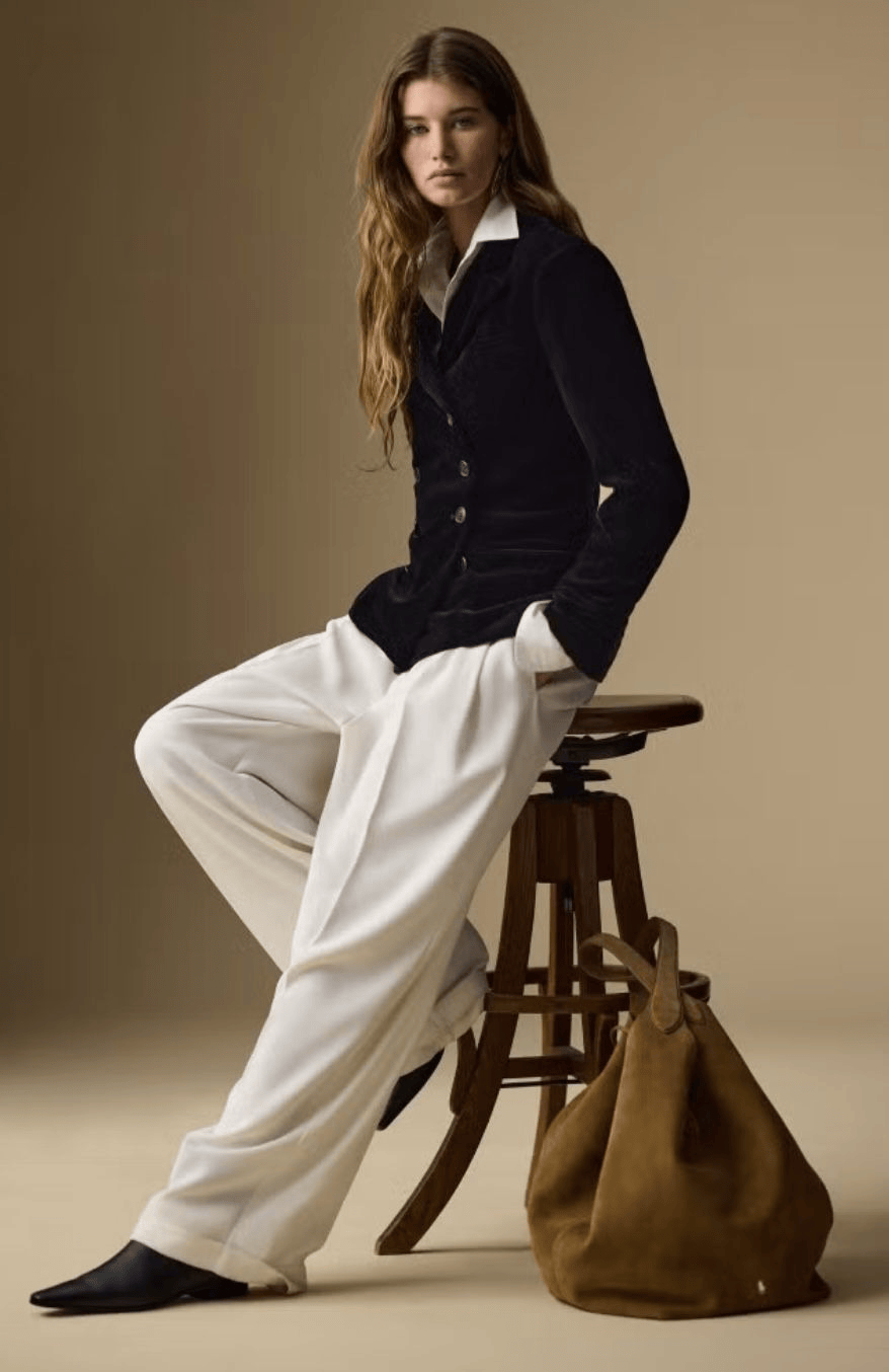 A woman with long wavy hair sitting on a wooden stool, wearing a dark blazer and white wide-leg pants, with a large brown bag on the floor next to her.