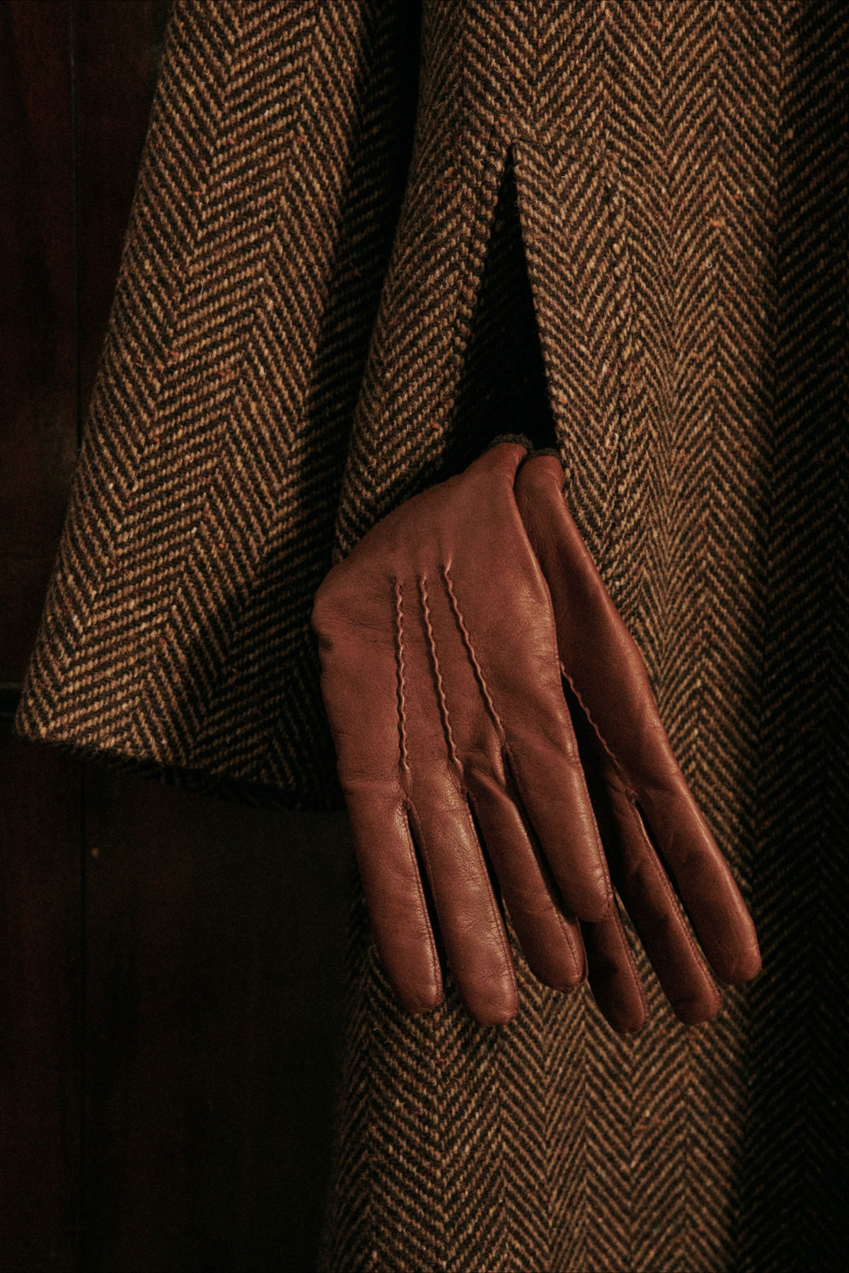 Close-up of a brown leather glove worn on the left hand, partially tucked into a garment made of brown and black herringbone patterned fabric.