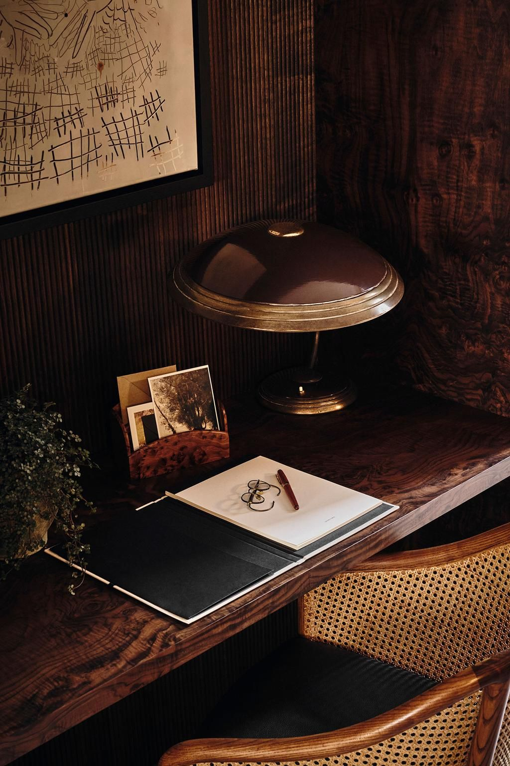 A dark wooden desk with a brass desk lamp, a pair of glasses, a red pen, a sketchbook, and photographs, with a wooden framed chair with woven cane backrest in a cozy, dimly lit room.