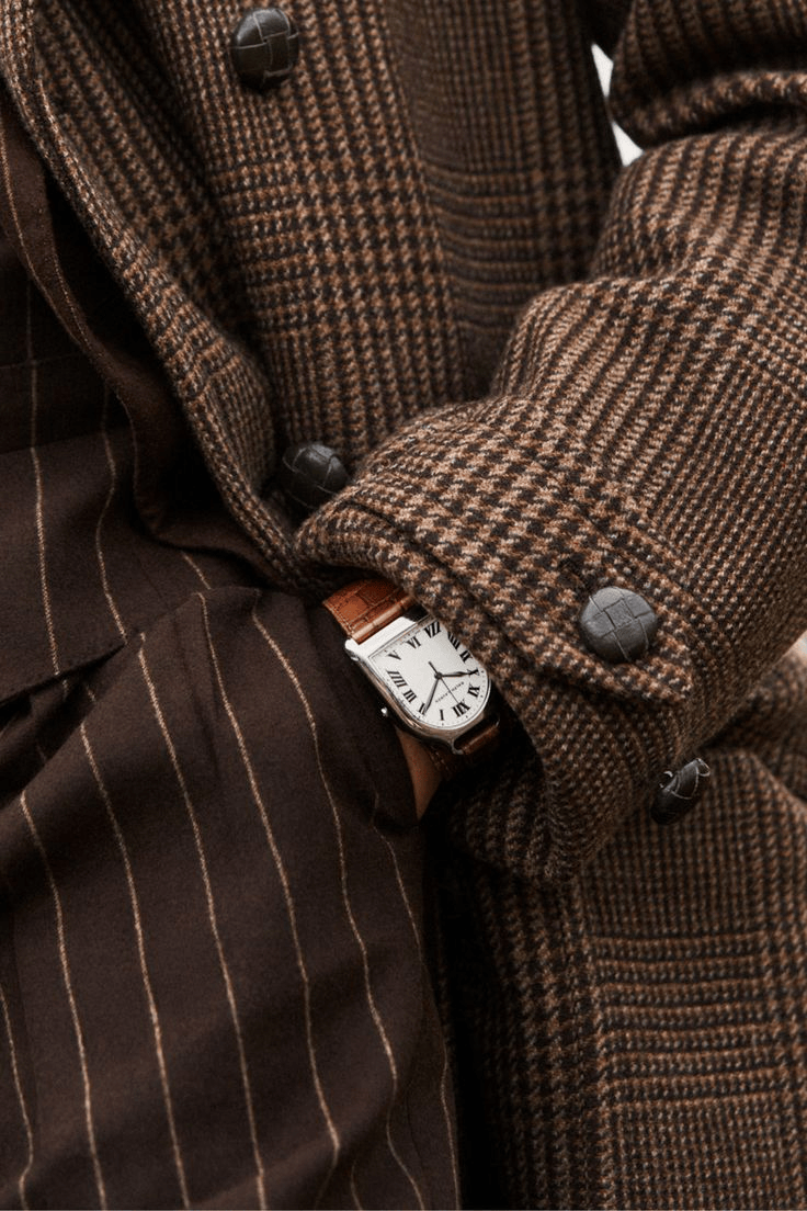 Person wearing a houndstooth patterned brown and black coat and pinstripe brown pants, with a wristwatch that has a white face and Roman numerals.
