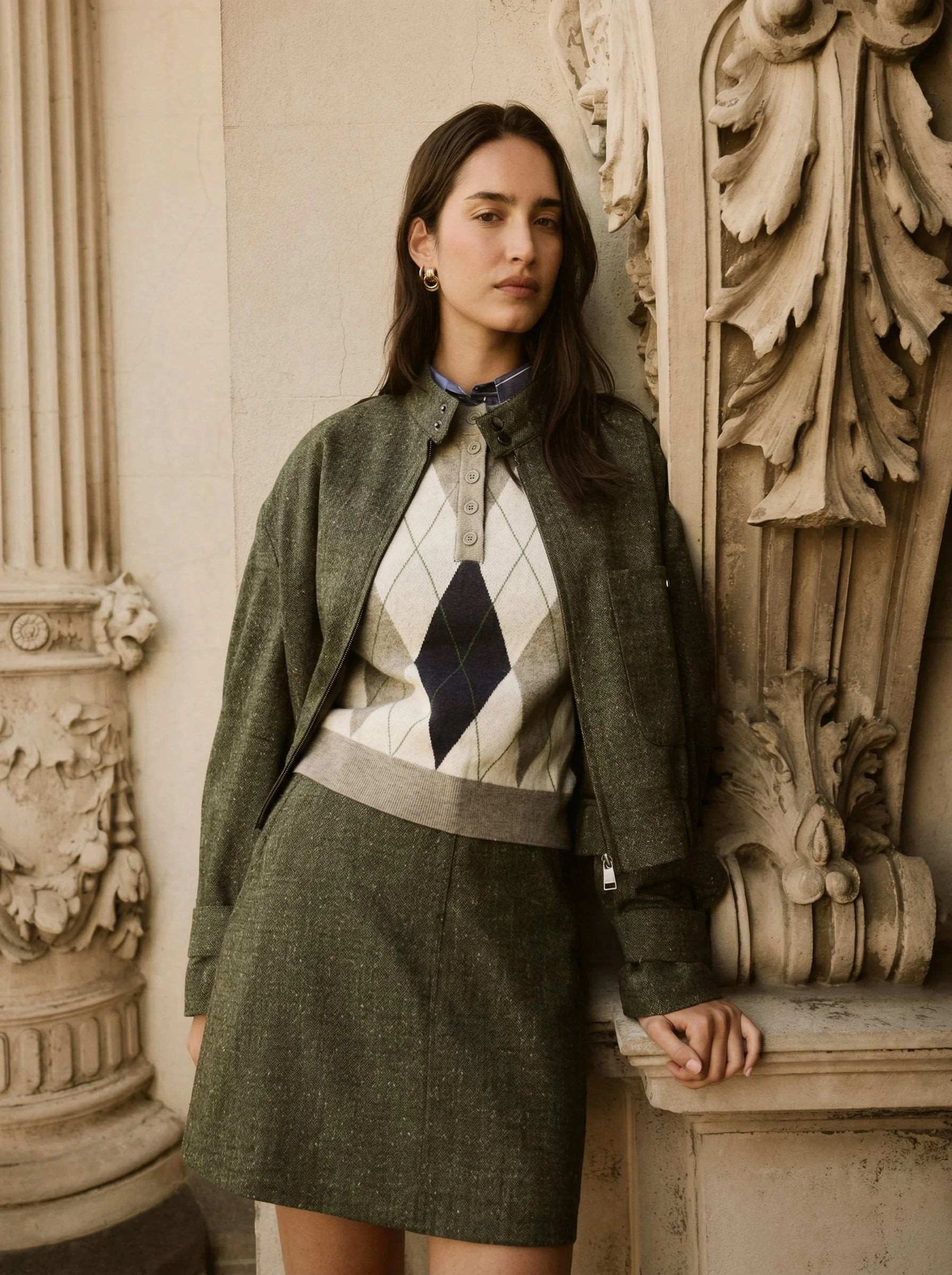 A woman with long brown hair wearing a green plaid skirt and matching jacket, with a beige and argyle sweater, standing next to an ornate stone wall with classical architectural details.