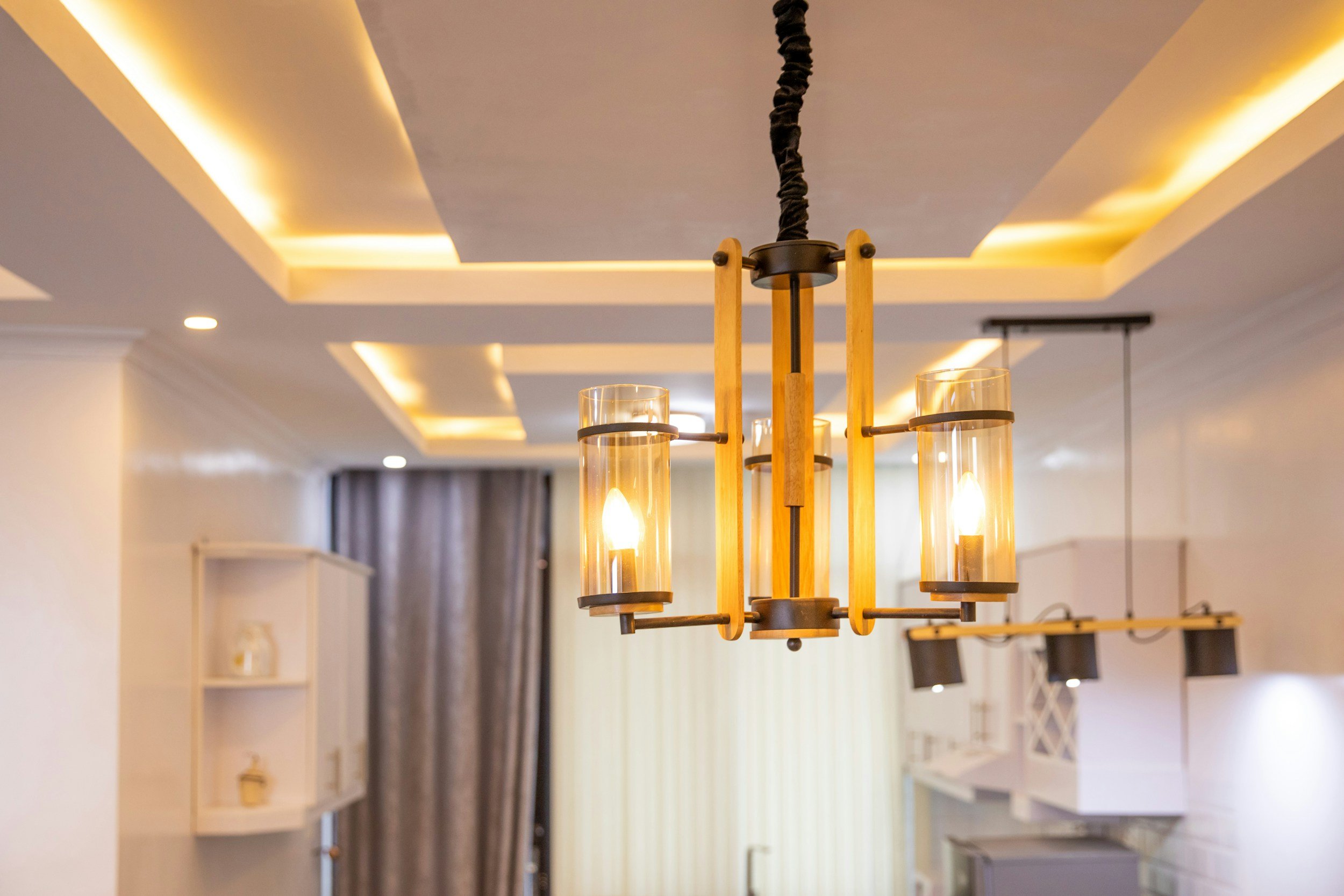 A modern chandelier with four exposed bulbs encased in cylindrical glass shades, hanging from a black twisted cord in a well-lit room with recessed ceiling lighting and a coffered ceiling design.