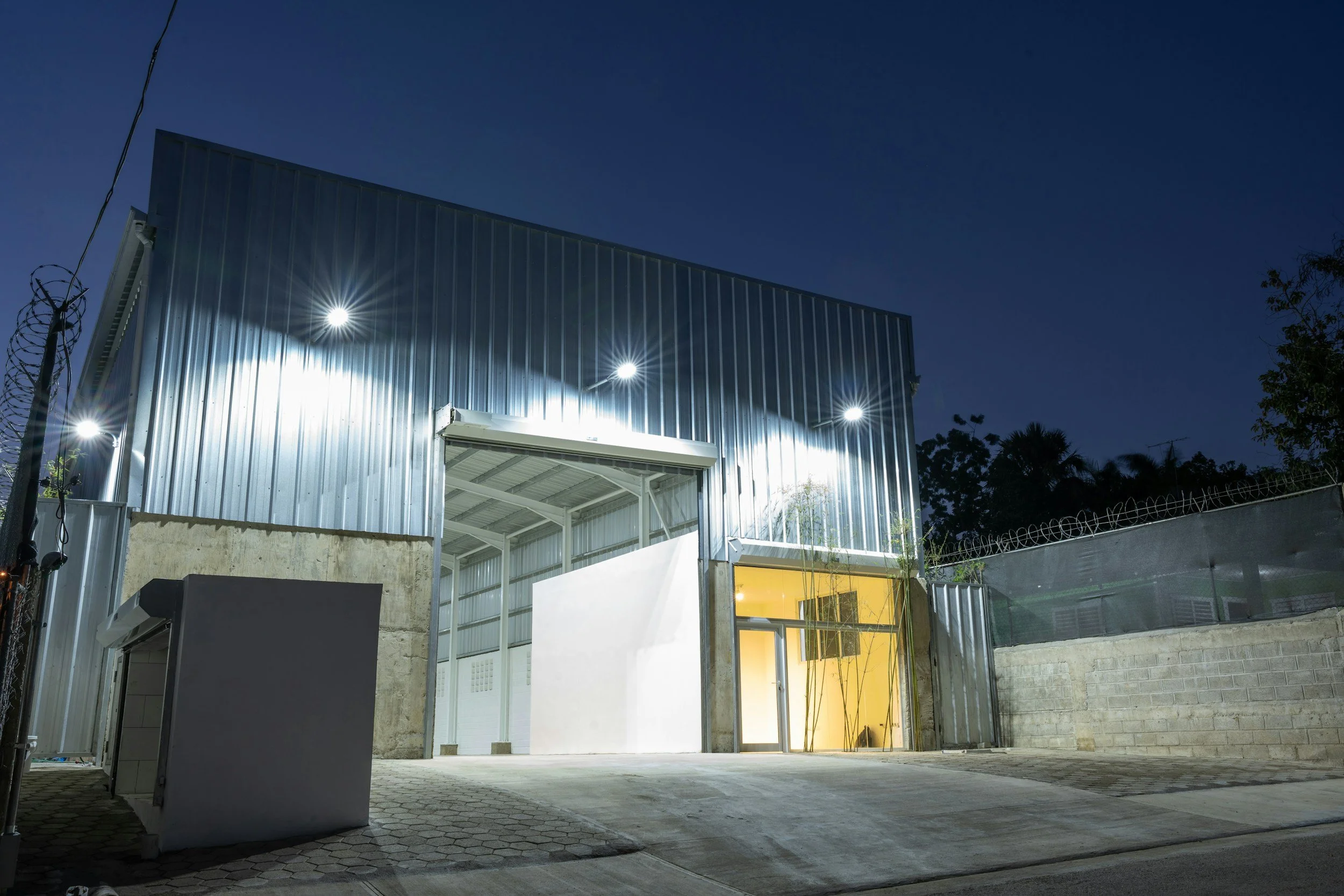 A modern industrial building at night with metal siding, bright exterior lighting, a yellow interior wall visible through the entrance, and a concrete driveway in front.