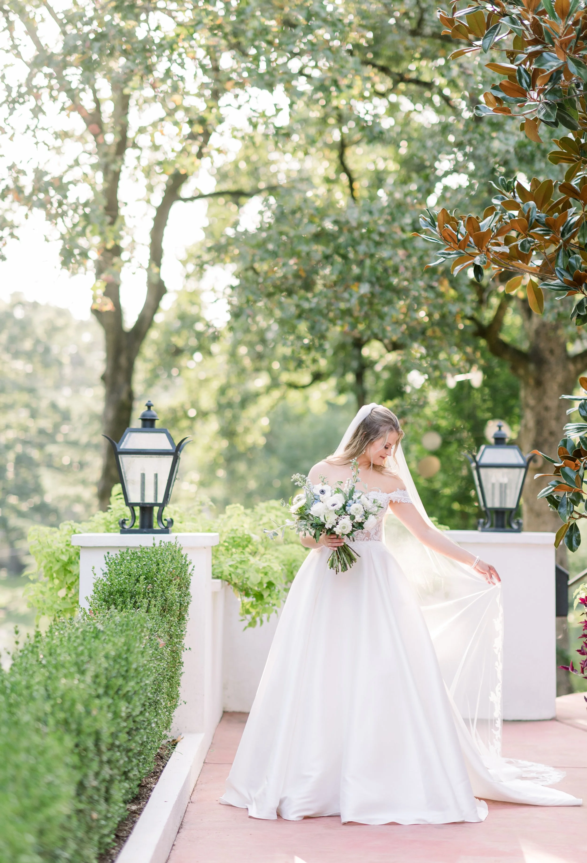 Bride in a white wedding dress holding a bouquet of white and purple flowers outdoors on a sunny day, standing on a pathway with greenery and trees in the background.