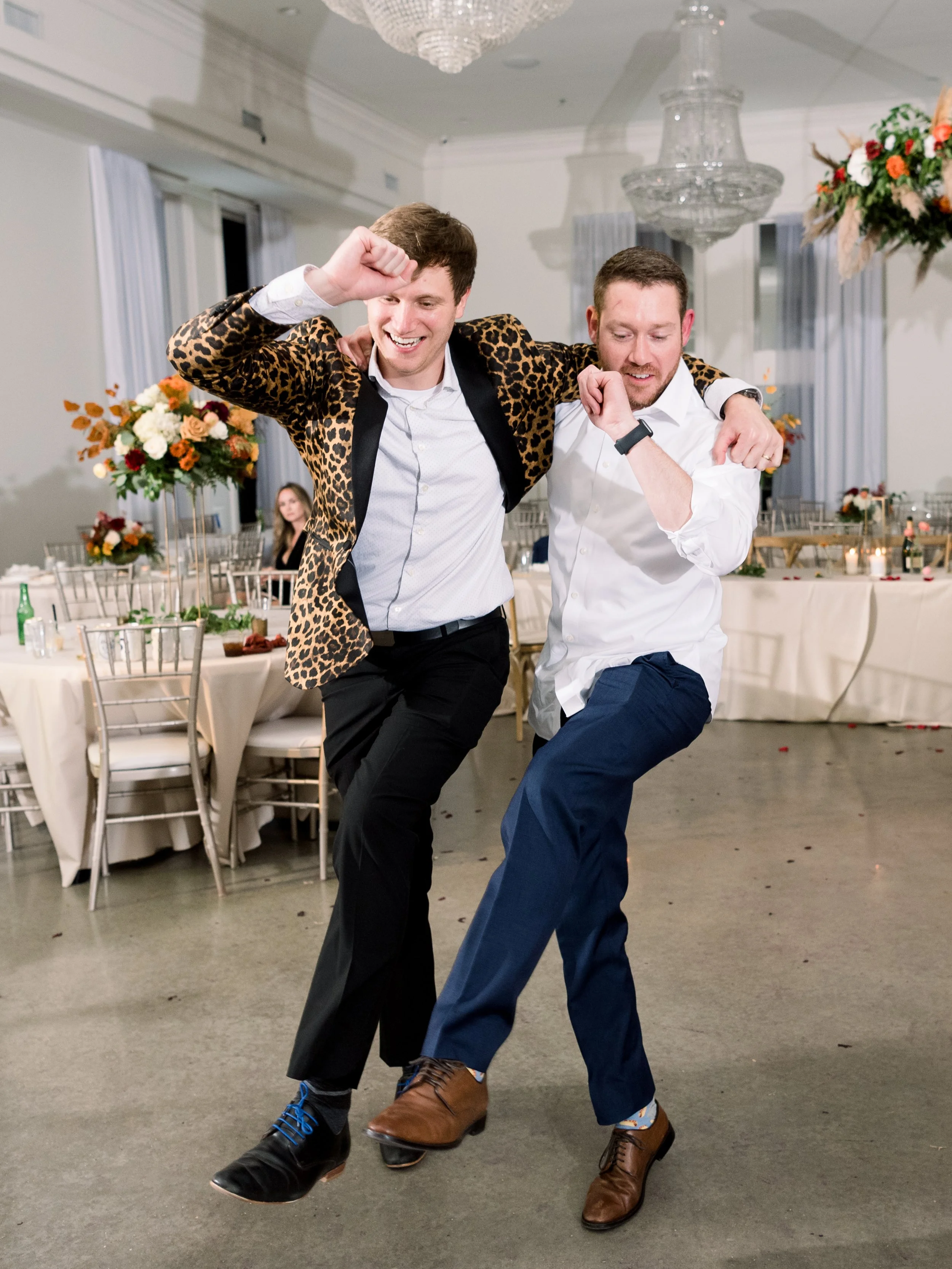 Two men dancing and celebrating at a wedding reception, one wearing a leopard print blazer and the other in a white shirt, with decorated tables and floral arrangements in the background.
