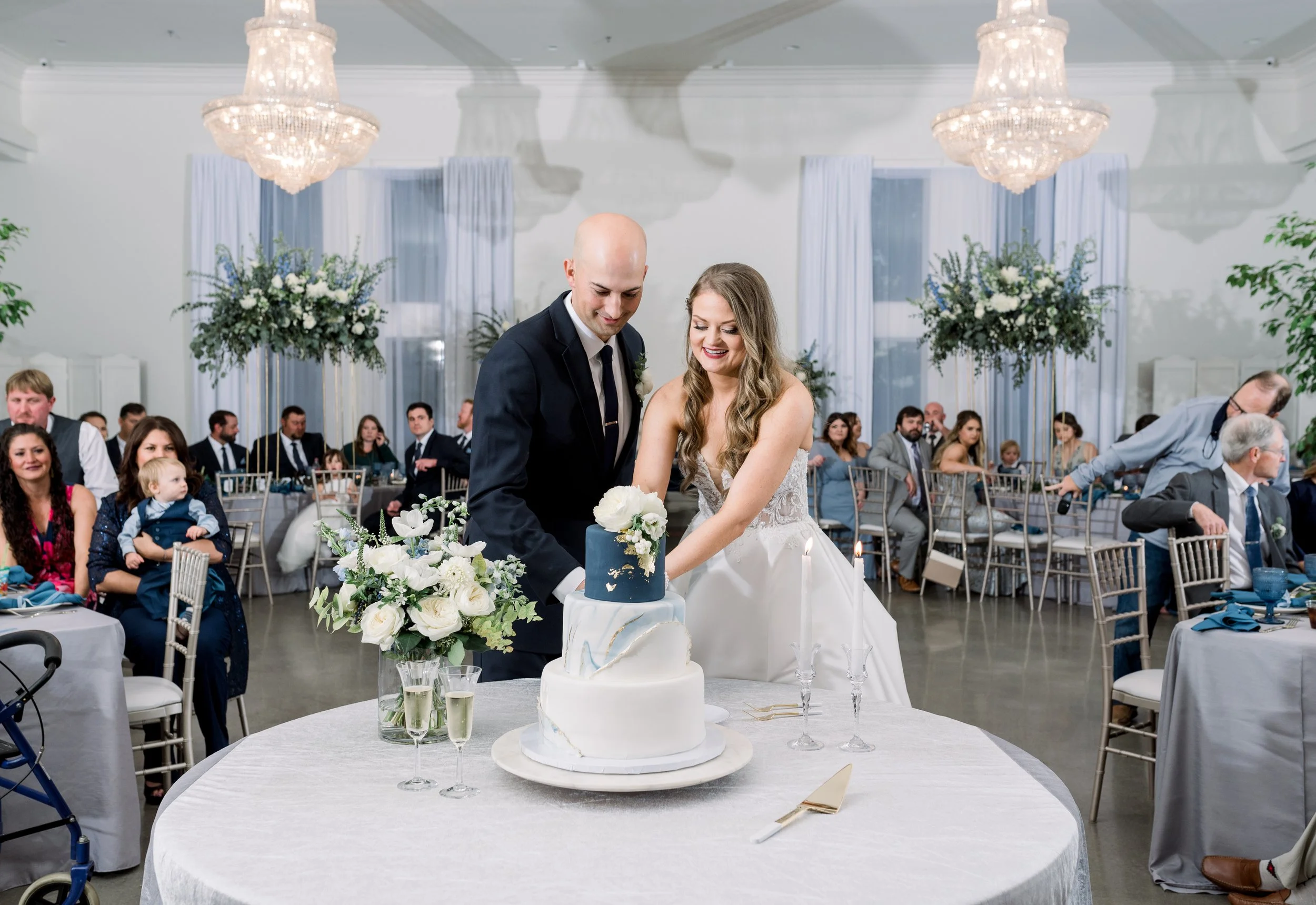 A bride and groom cutting a wedding cake at their wedding reception, surrounded by guests seated at decorated tables with floral arrangements.