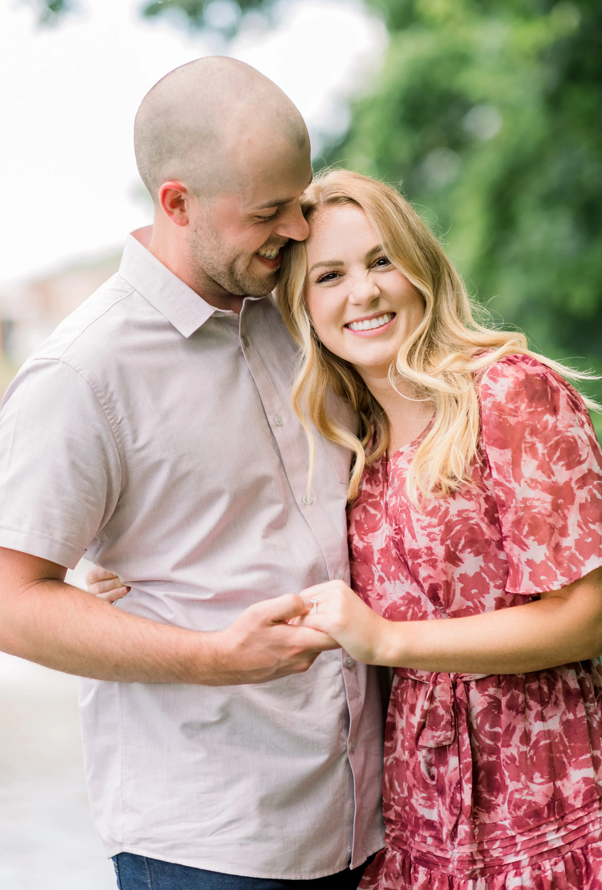 A smiling couple holding hands outdoors, with the man leaning his head toward the woman's forehead, and the woman winking.