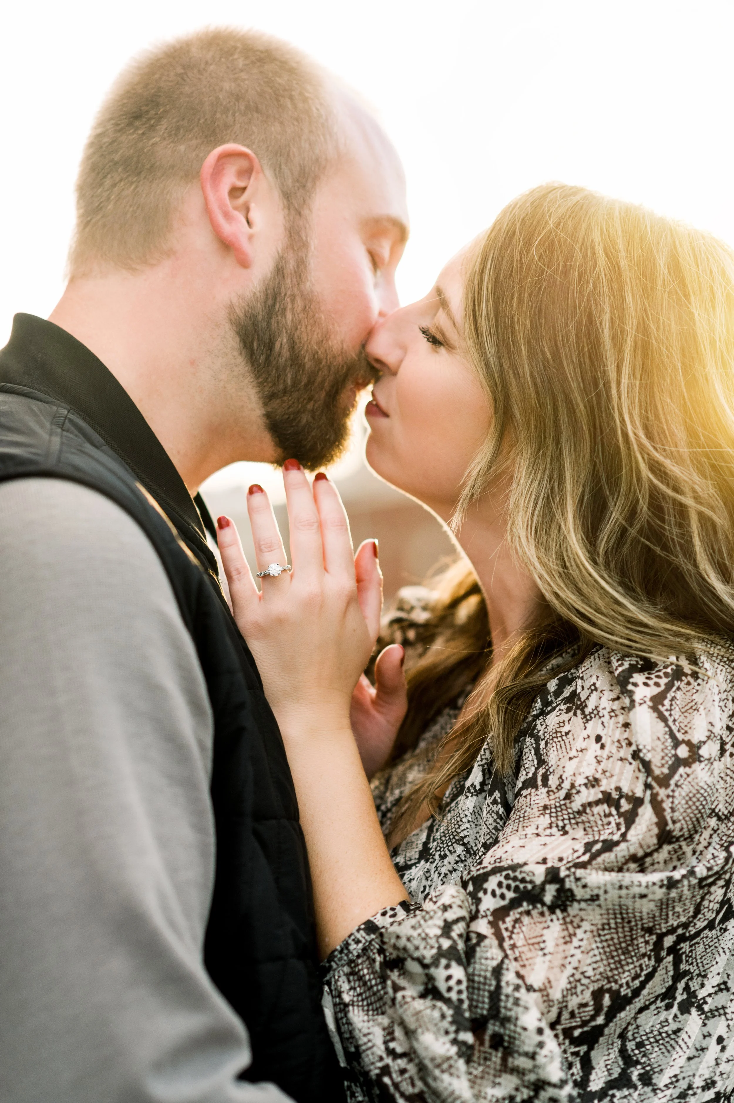 A couple sharing an intimate kiss, with the woman wearing an engagement ring, and sunlight shining on their faces.
