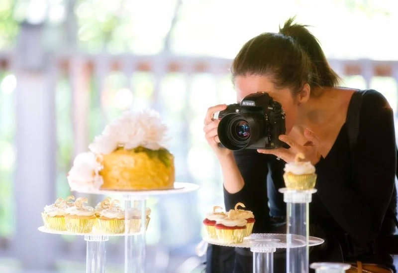 A woman taking a photo of cupcakes and a cake with a camera