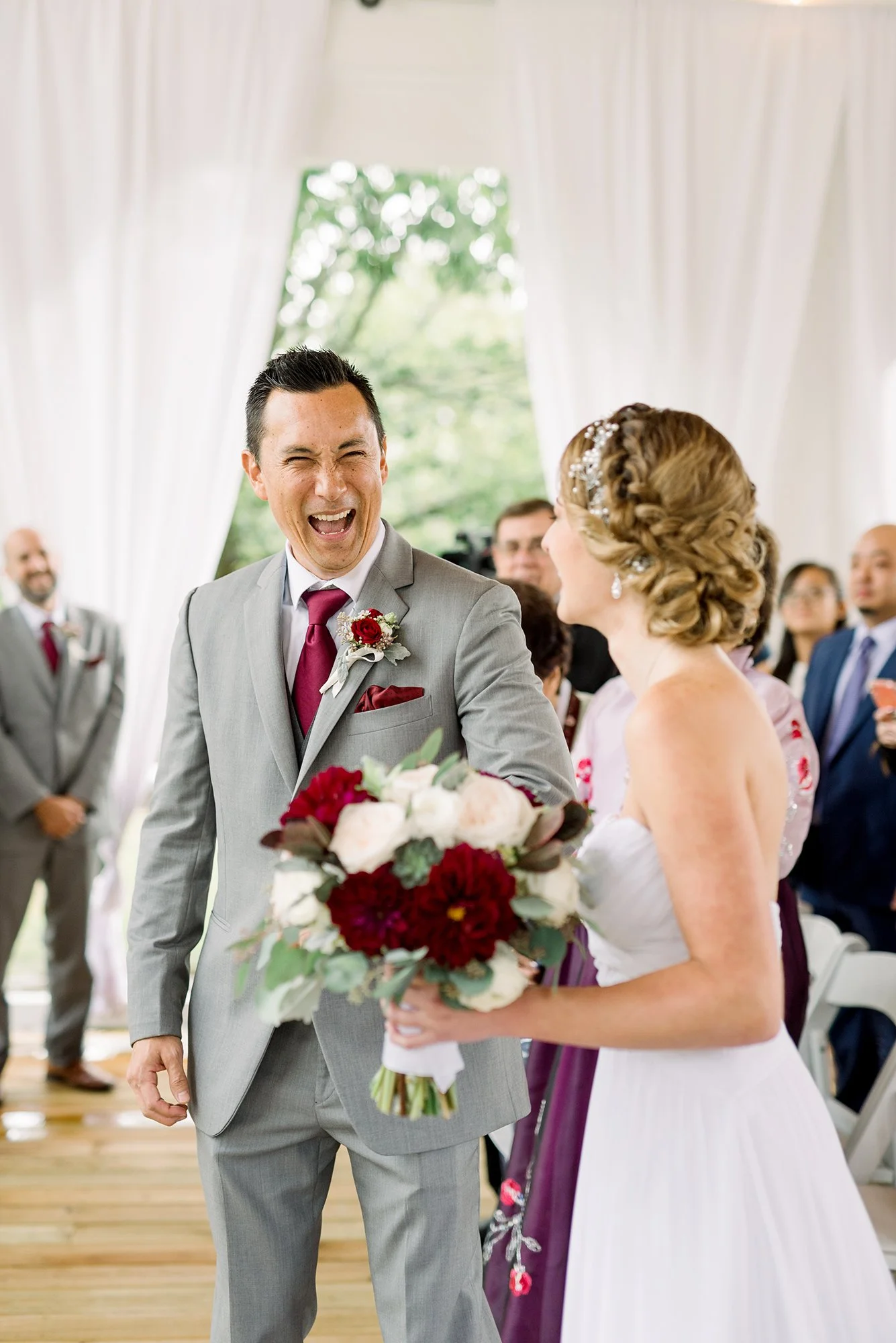 A joyful groom in a gray suit with a red tie and boutonniere at wedding, smiling and looking at the bride, who is holding a bouquet, in a bright venue with dressed guests in the background.