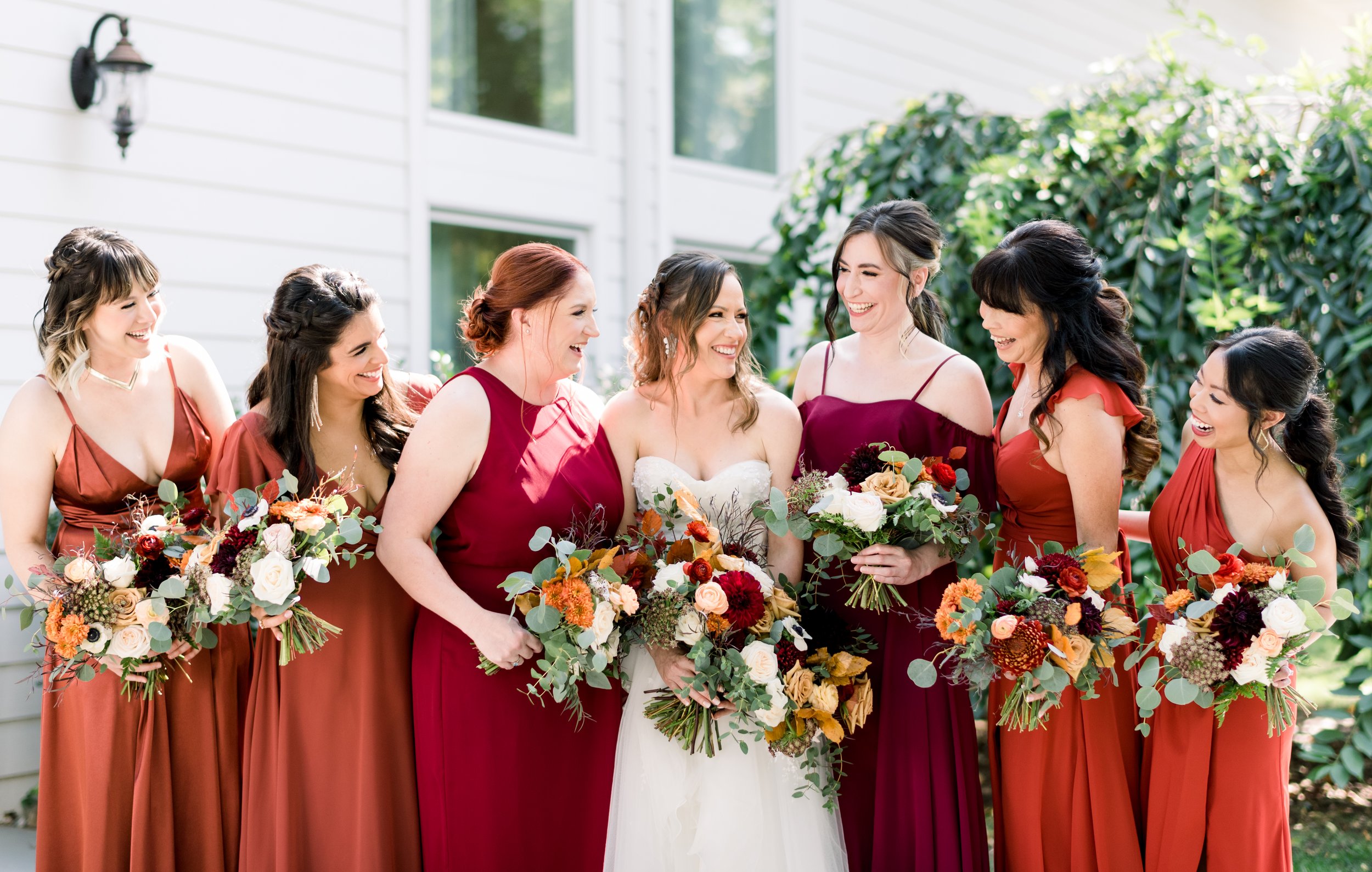 A bride and seven bridesmaids standing outdoors, smiling and holding floral bouquets, with a white house and greenery in the background.