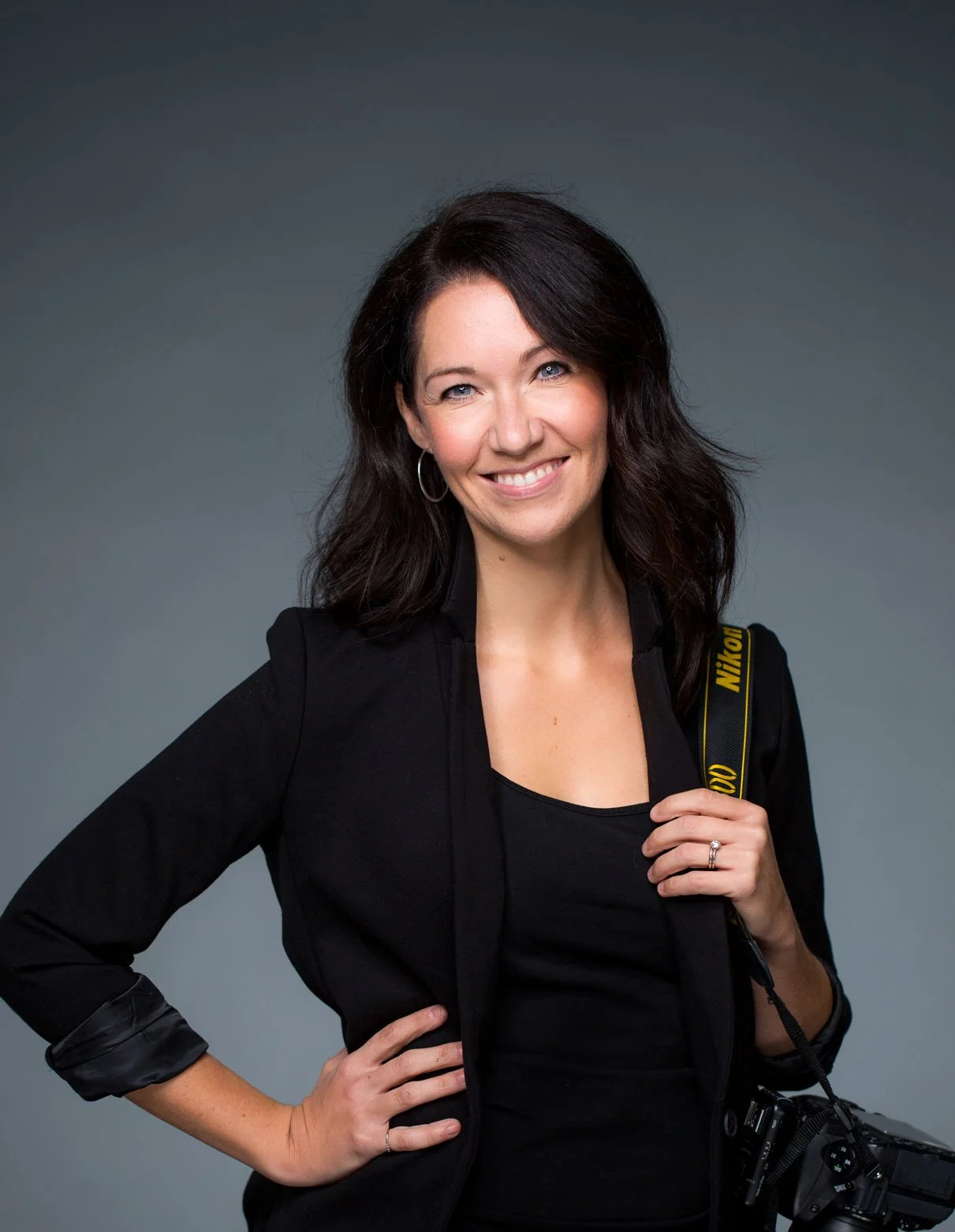 A woman with dark hair, wearing a black outfit and holding a Nikon camera strap over her shoulder, smiling and standing against a gray background.