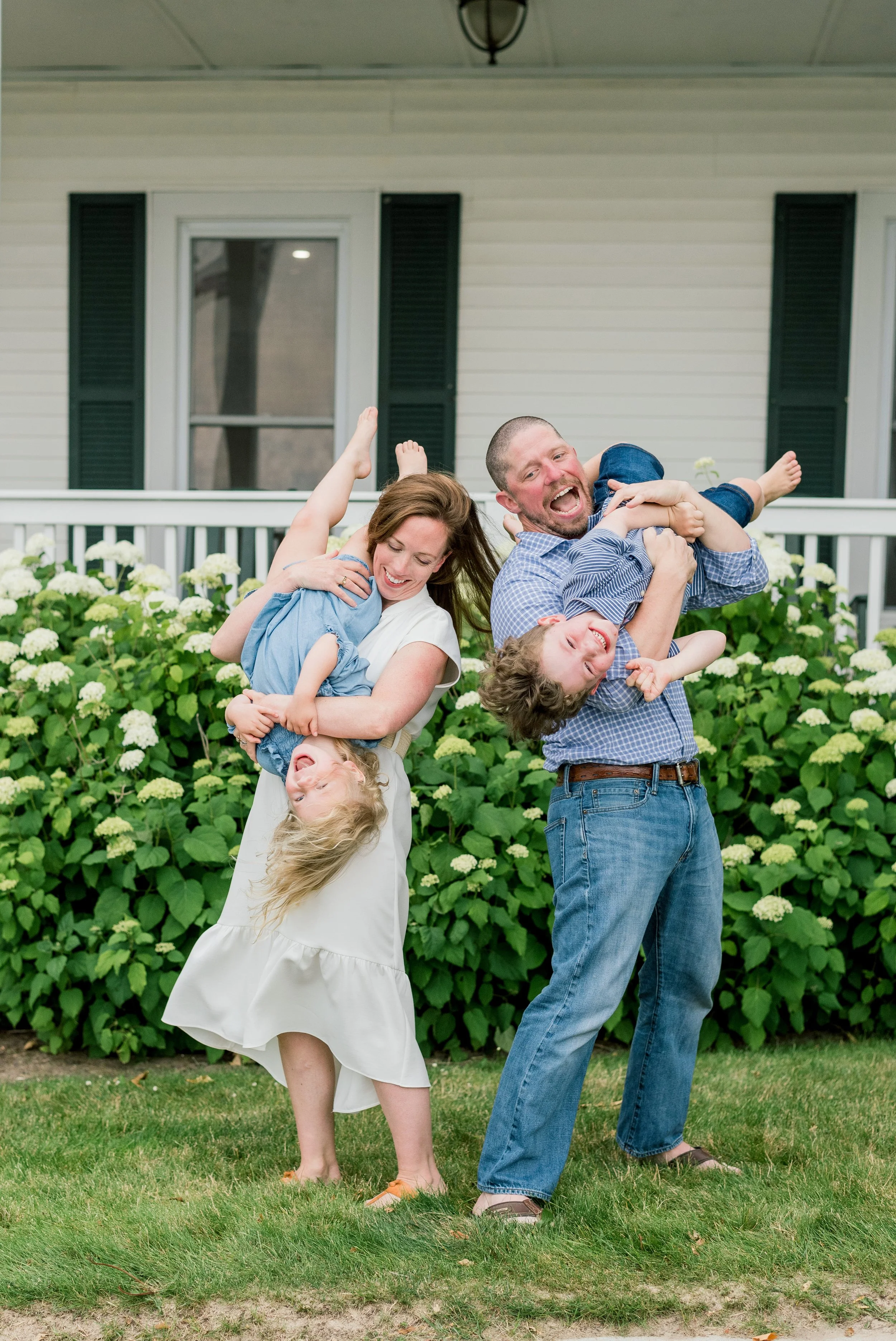 Family of four playing and laughing outdoors in front of a house, with two parents lifting their children, a young girl and boy, into the air, all smiling and enjoying the moment.