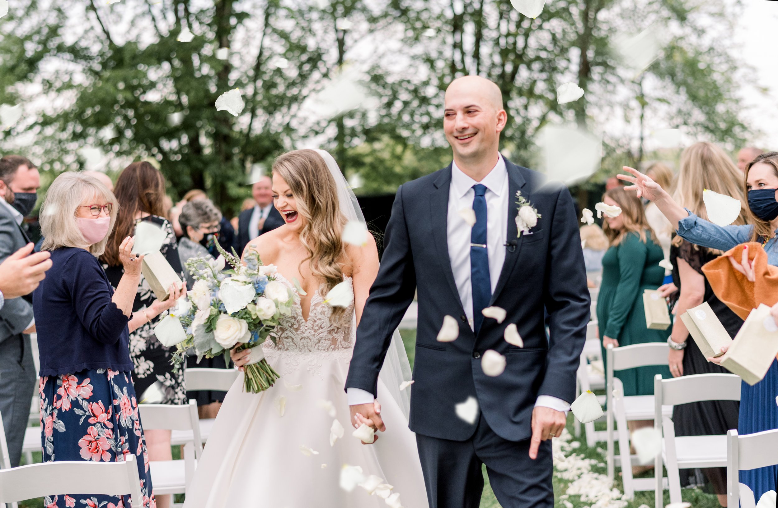 Bride and groom walking hand in hand down an outdoor wedding aisle, smiling, surrounded by guests throwing flower petals.