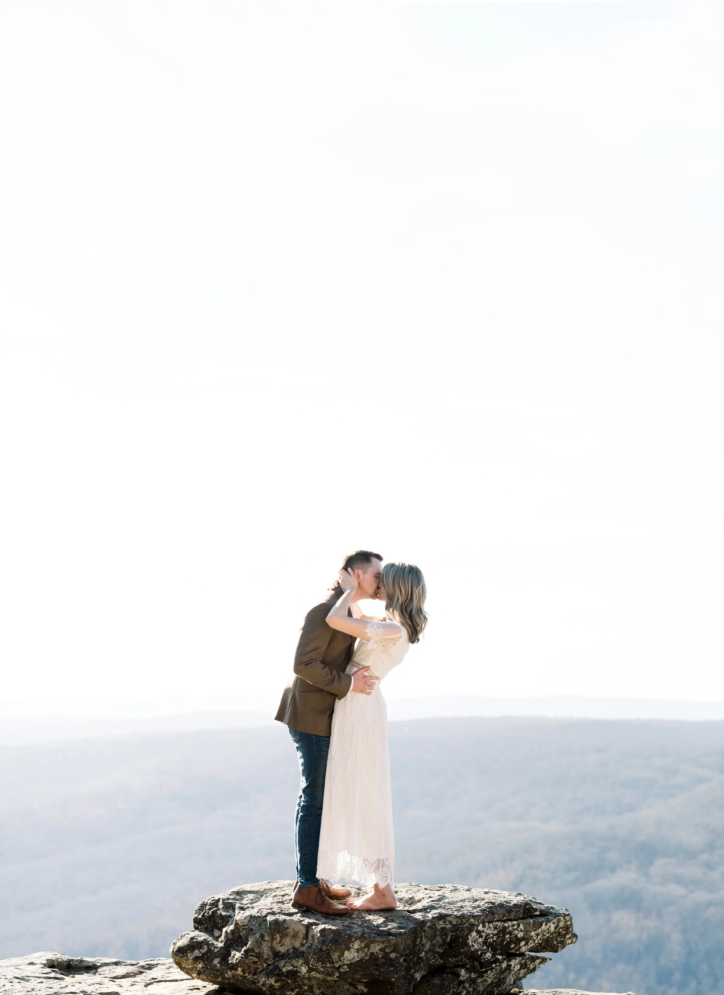 A couple stands on a large rock at the edge of a cliff, embracing and kissing against a bright sky background, with a scenic mountain landscape in the distance.