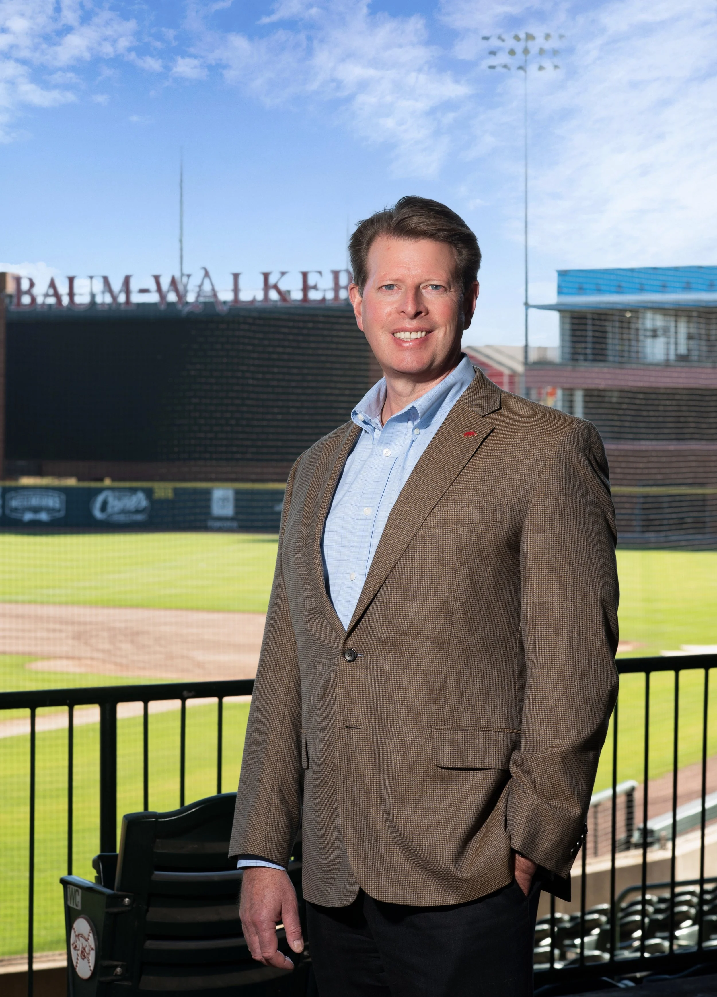 A man in a brown blazer and light blue shirt standing at a baseball stadium with a field and scoreboard in the background.