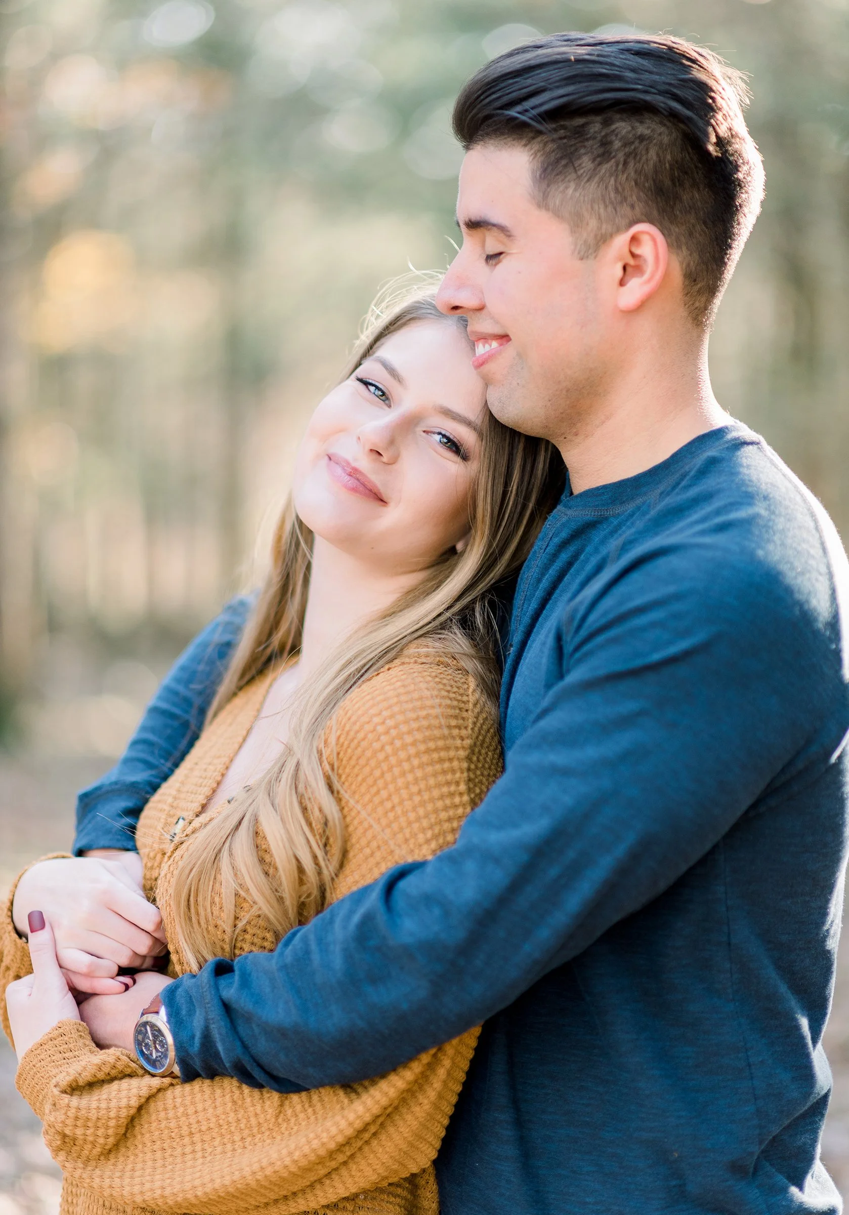 A young couple standing closely outdoors, with the woman leaning her head on the man's shoulder. Both are smiling softly, with the woman wrapping her arms around his waist. The background is blurred trees, indicating a natural setting during daylight