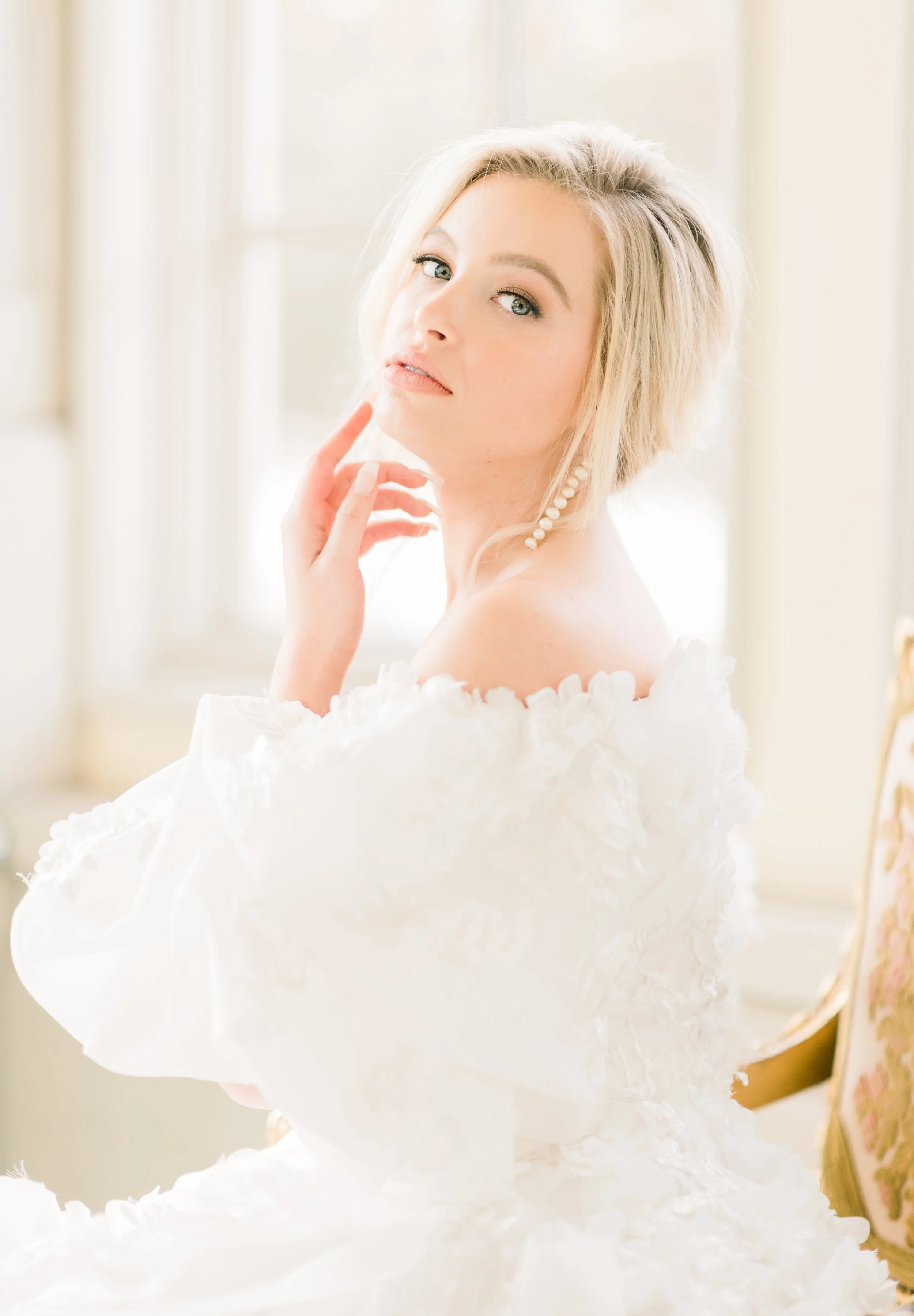 A young woman with blonde hair, blue eyes, and light makeup, wearing a white off-shoulder dress with ruffled details and pearl earrings, touching her face and looking at the camera, posed in a well-lit indoor setting.