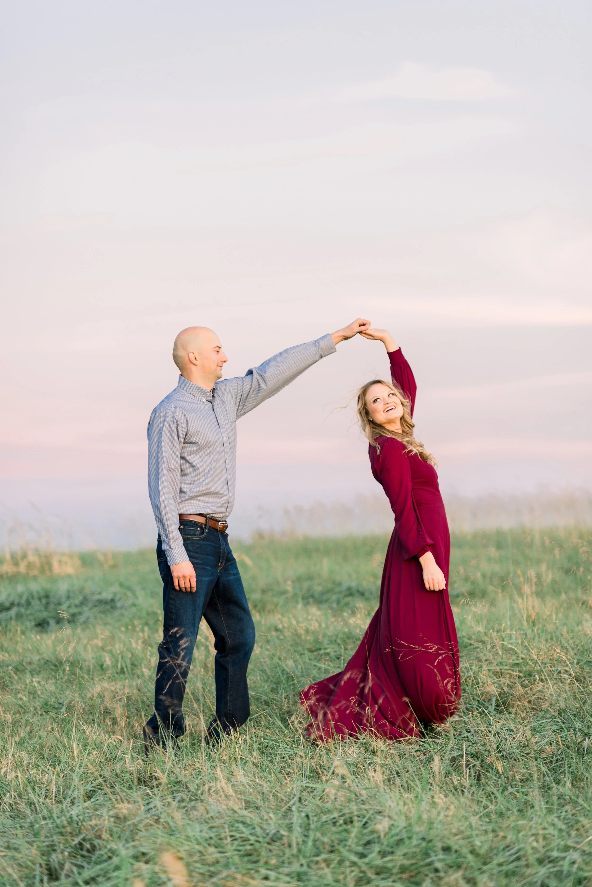 A man and a woman dance in a grassy field at sunset, holding hands as he twirls her in a flowing burgundy dress.