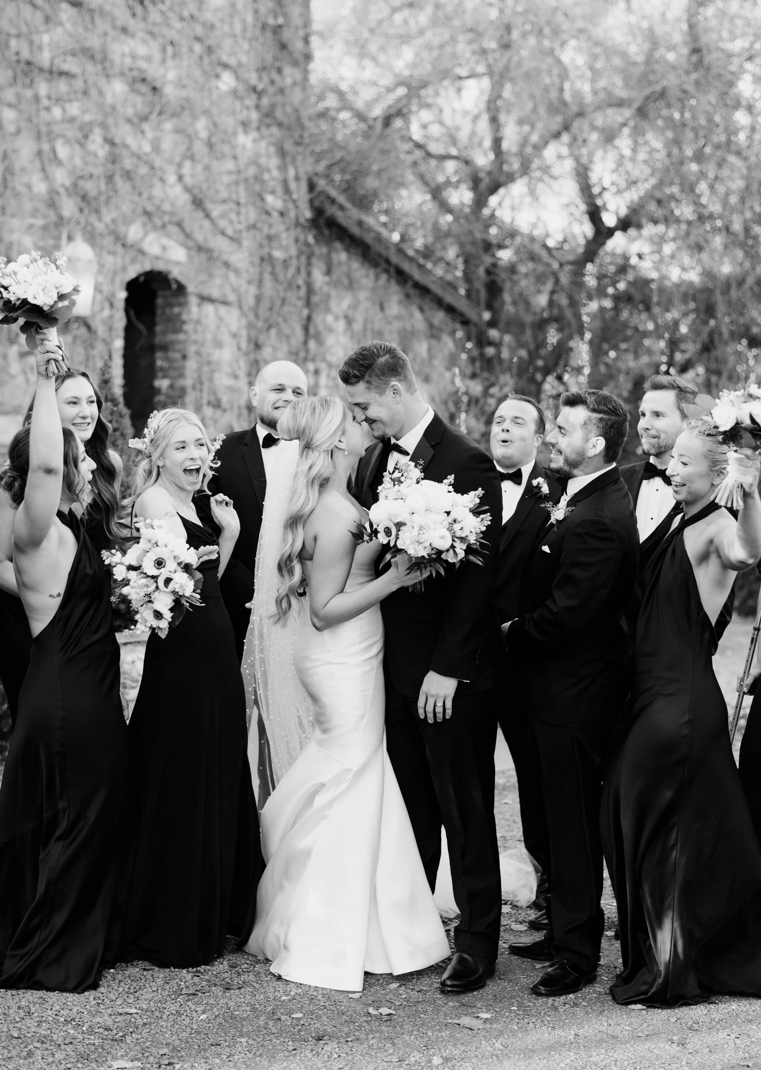 A black and white photo of a wedding celebration outdoors, featuring a bride and groom embracing and exchanging vows, surrounded by bridesmaids and groomsmen smiling and holding bouquets.