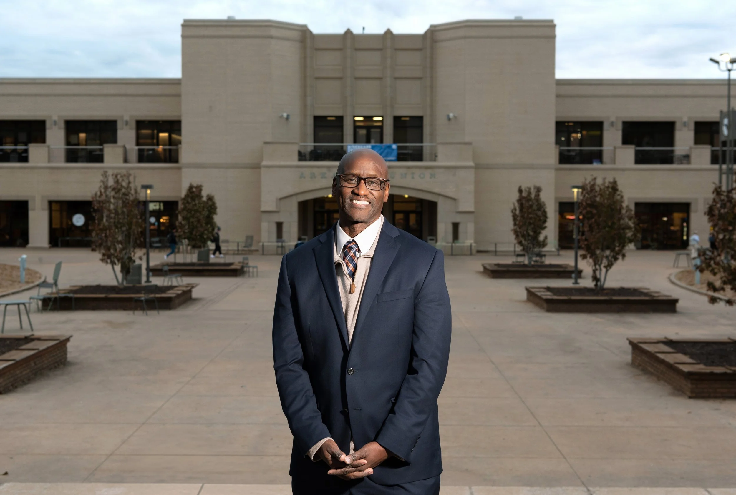 A smiling man wearing glasses, a dark suit, and a tie, standing outside in front of a public building.