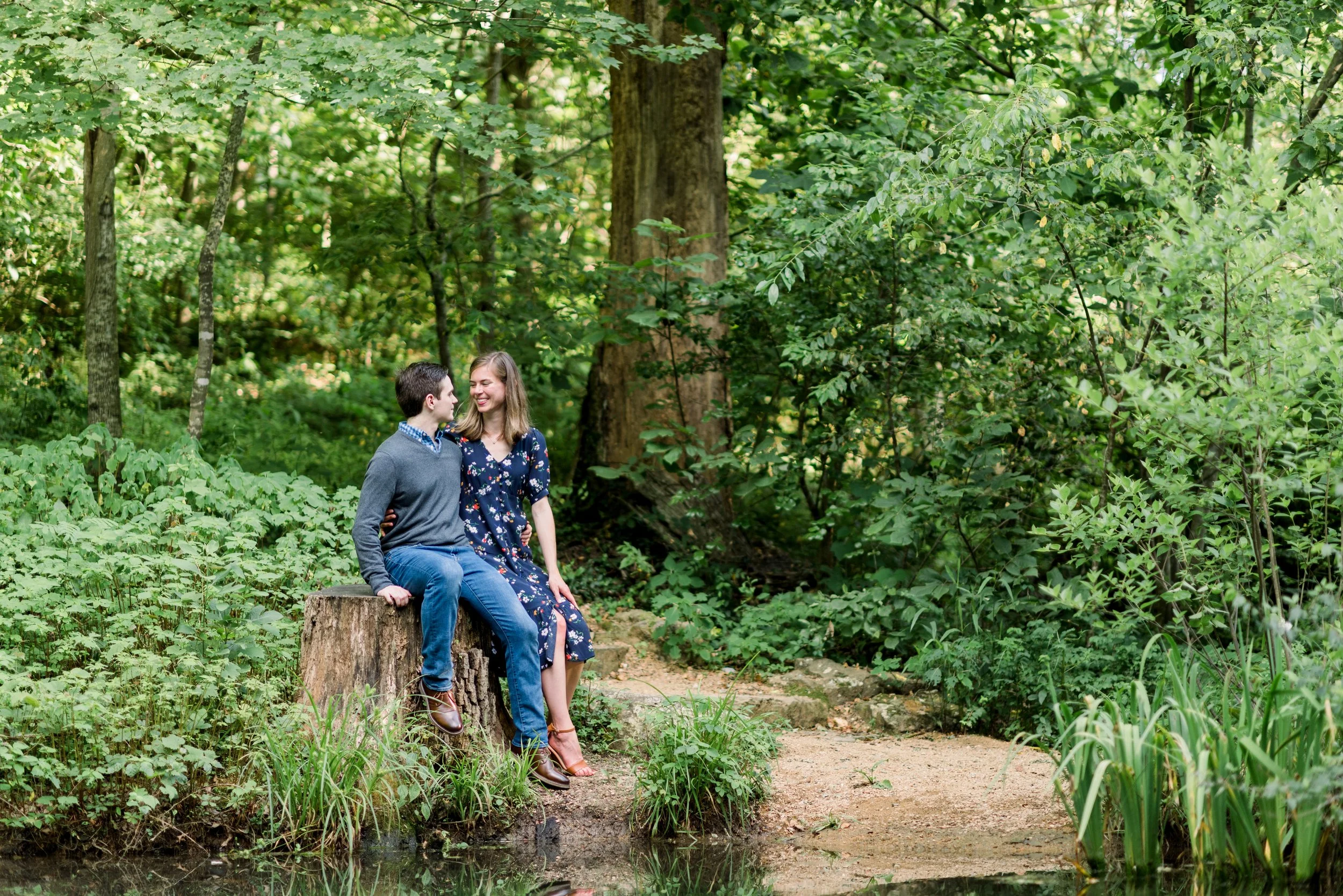 A young man and woman sitting on a tree stump in a lush green forest, smiling and looking at each other.