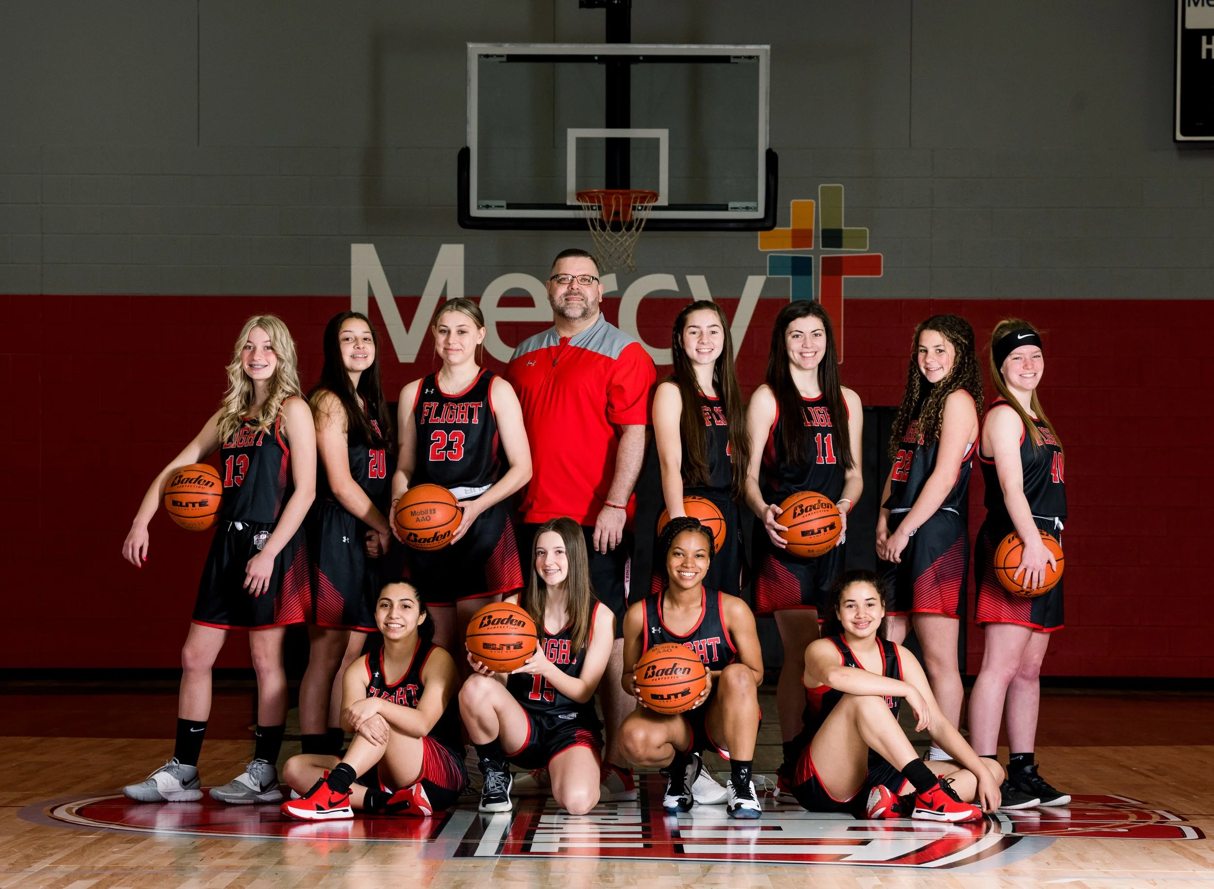 A girls' basketball team posing in a gymnasium in front of a wall with the Mercy logo. The team members are holding basketballs and are wearing matching black and red jerseys. The coach stands behind them.