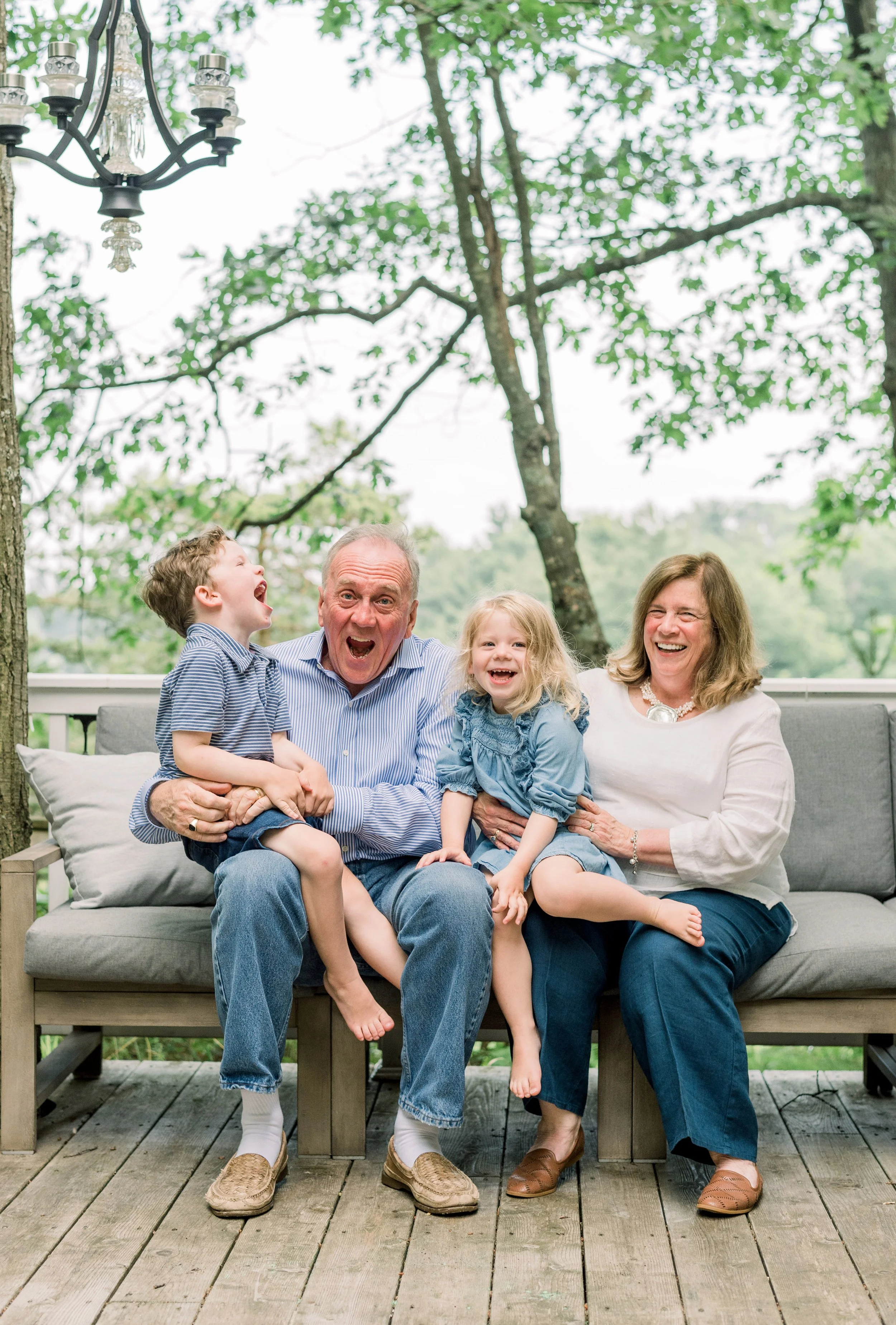 A happy family of four sitting on a gray outdoor couch on a wooden deck, surrounded by lush green trees, laughing and enjoying each other's company.