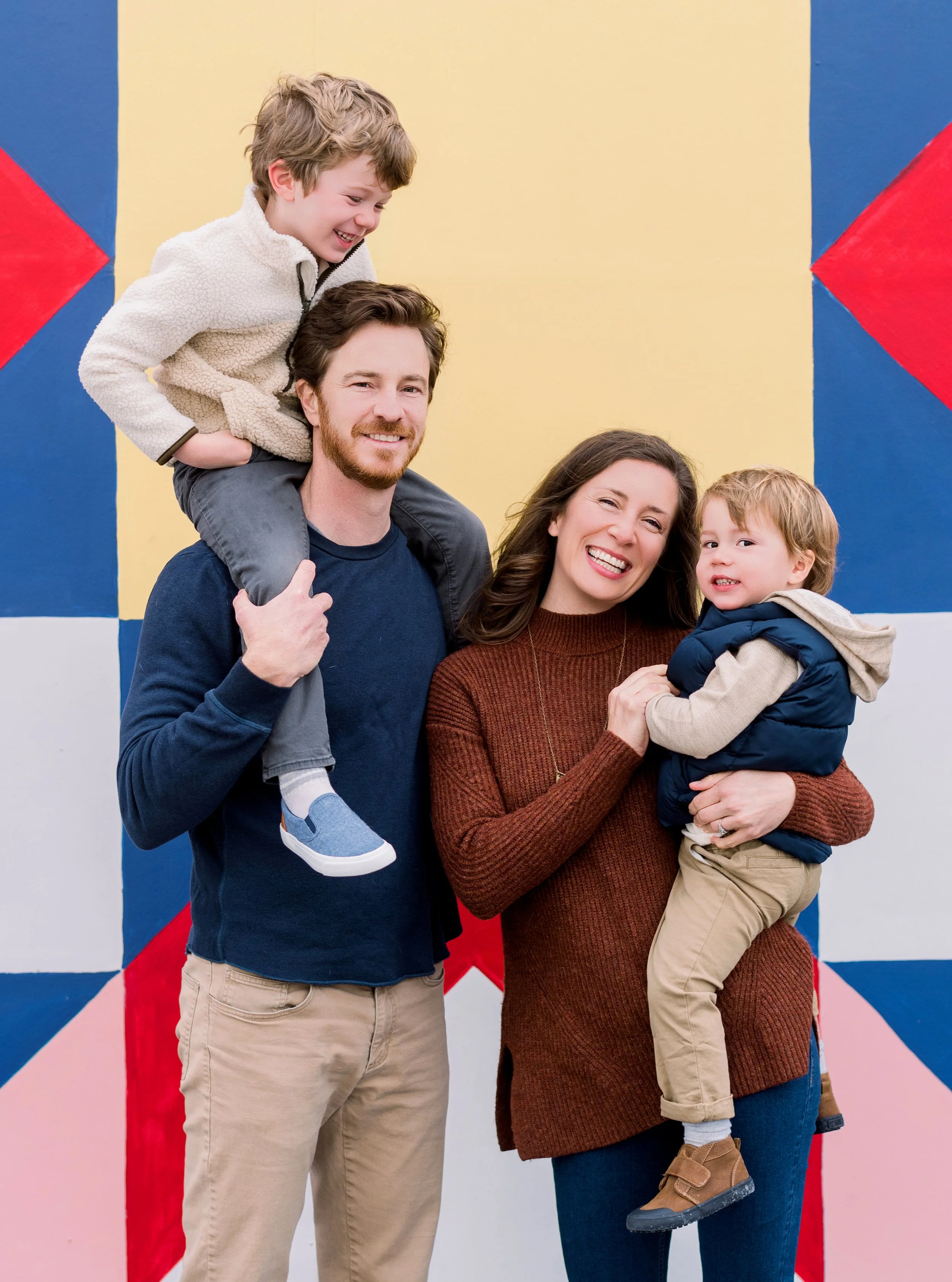 A happy family of four outdoors, two adults and two children, standing in front of a colorful geometric mural. The man has a boy on his shoulders, smiling, and the woman has a younger boy in her arms, both smiling.
